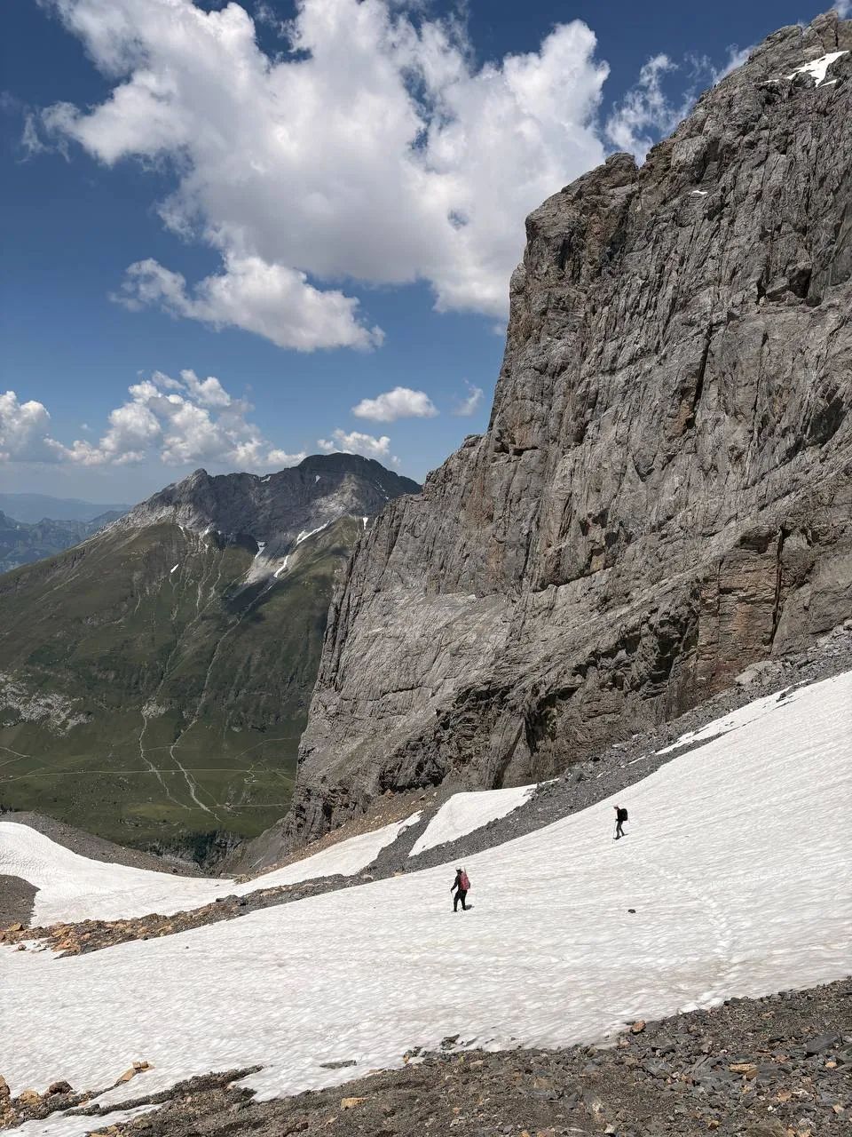 Two tiny figures crossing a snowfield beneath a towering rock wall, green valley far below.