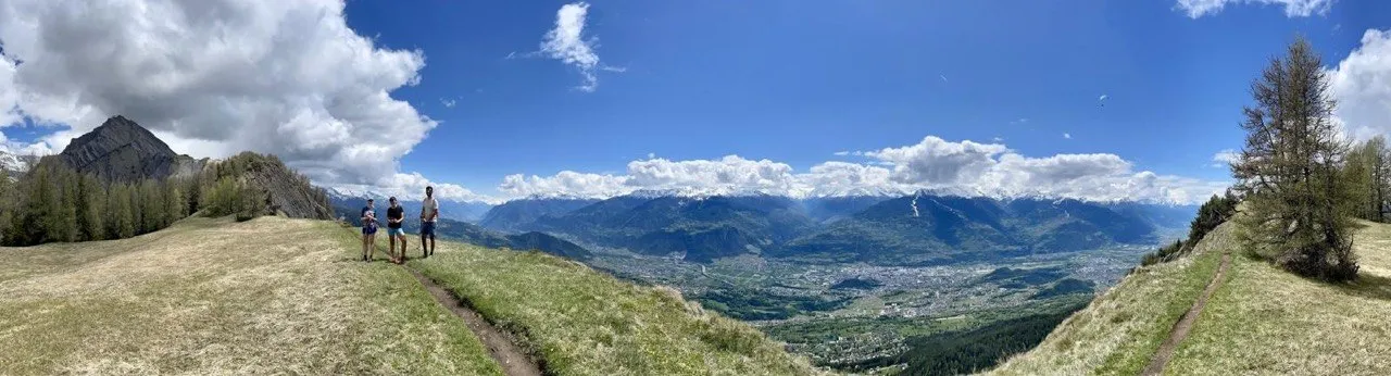 Panoramic view from Prabé over the Rhone valley, three hikers standing on the grassy summit.