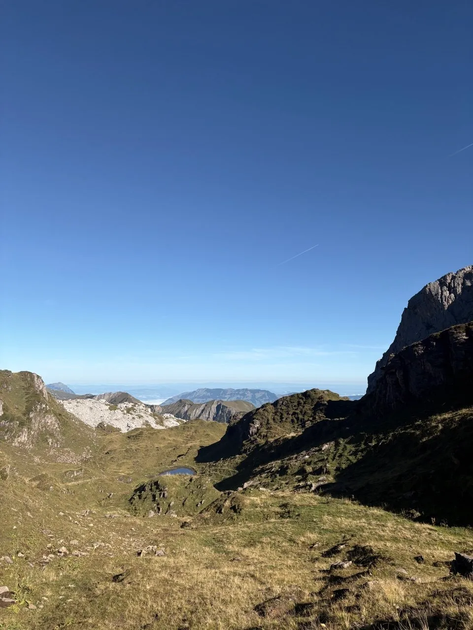 High basin with a small tarn and limestone karren, wide Alpine ranges under a blue sky behind.