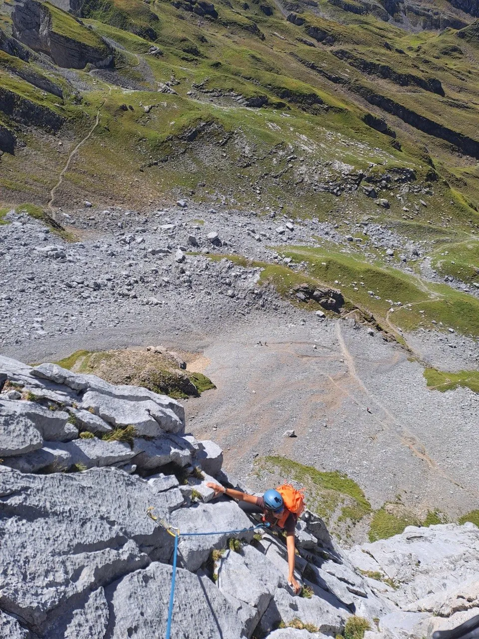 Looking down the wall at a climber on the rope, scree slopes, green hillsides and the approach path far beneath.