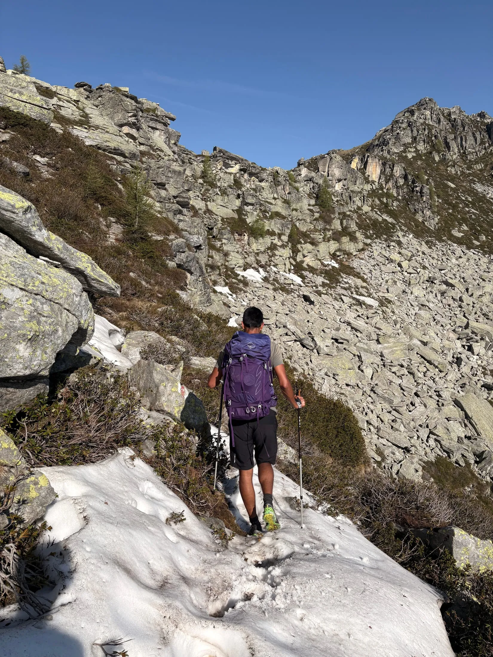 A hiker with poles crosses a small snowfield on a rocky slope under a clear sky.