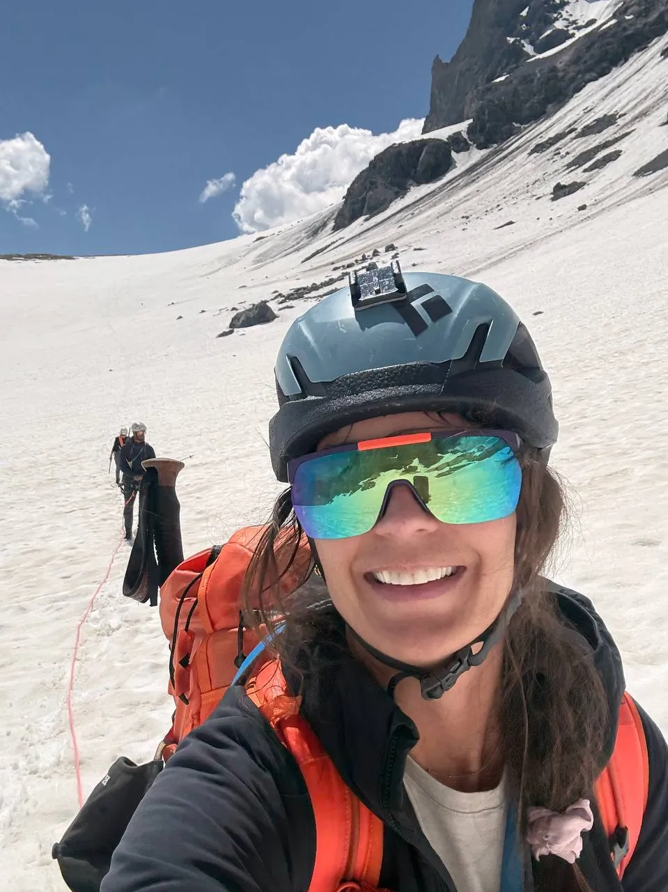 Selfie in helmet and sunglasses on the snow, a roped companion coiling the rope behind.