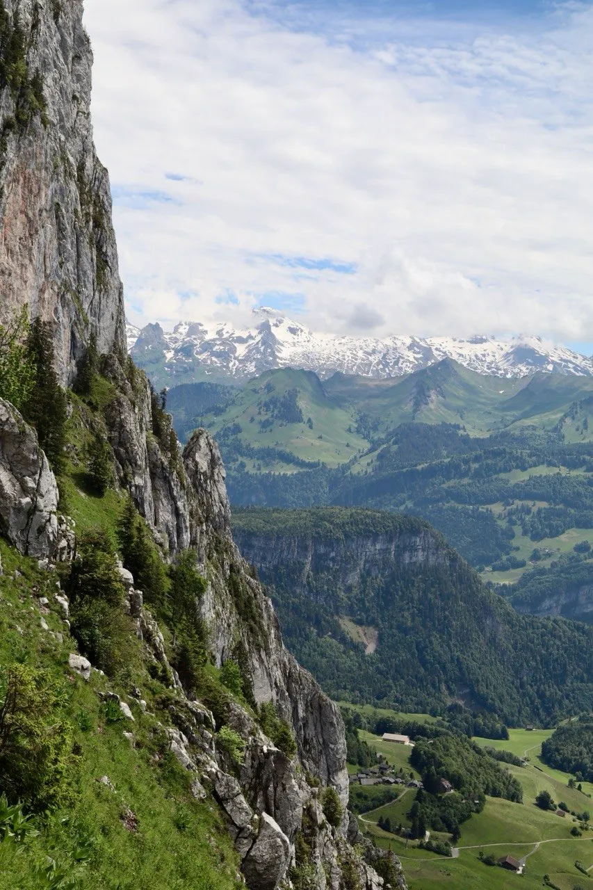 Green valley and snow-capped peaks framed by a towering limestone cliff.