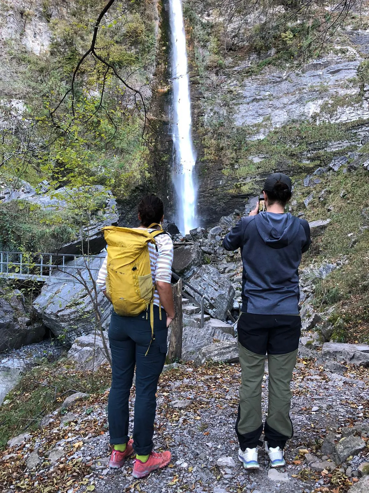 Two hikers watching a tall thin waterfall drop down a mossy rock face.