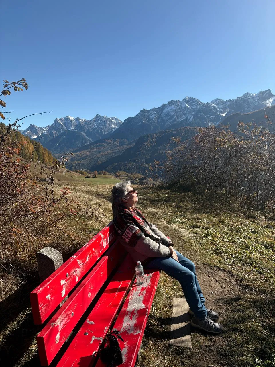 My mother sitting on a red bench in the November sun, looking across to the snowy peaks.