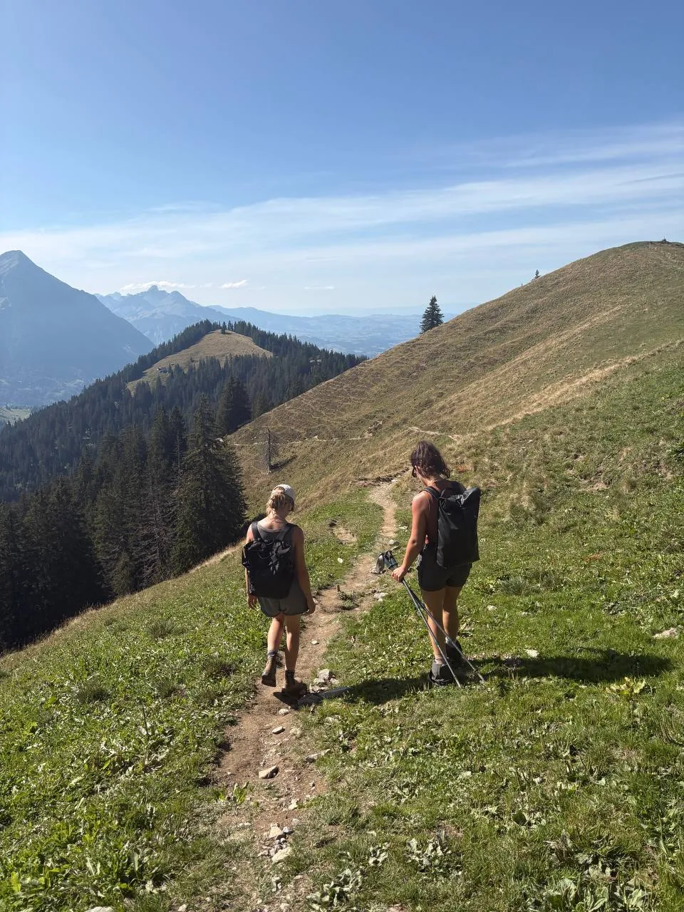Two hikers on a narrow path running out along a broad grassy ridge, forest and distant peaks all around.