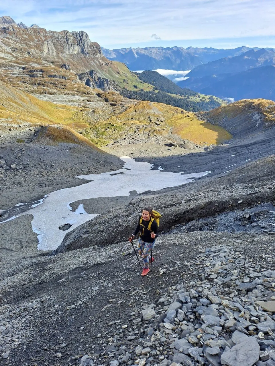 Hiker crossing a steep scree slope with patches of snow, wide valley below.