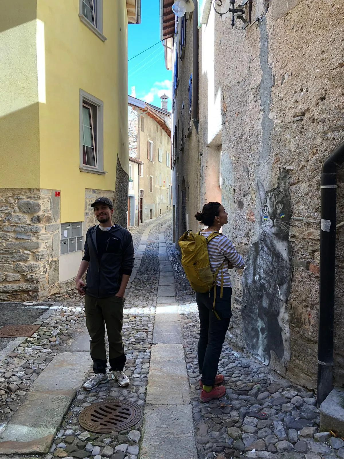 Two people in a narrow cobbled village alley beside a large cat mural.