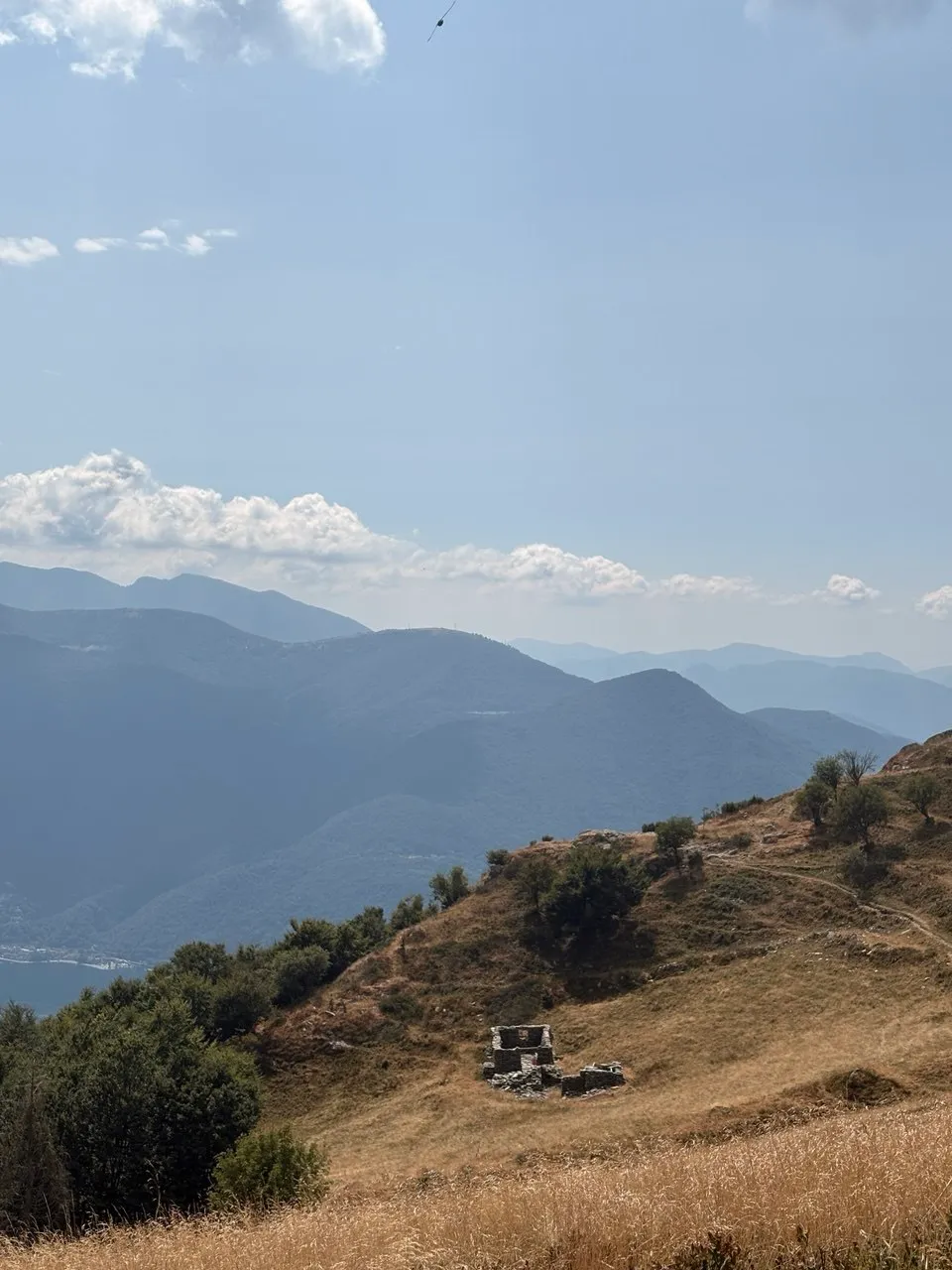 The remains of an old stone building on a dry grassy hillside, hazy blue ridges and a sliver of the lake beyond.