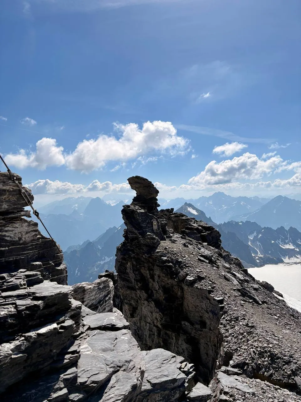 Rocky summit ridge with a fixed cable and a hazy panorama of snowy peaks beyond.