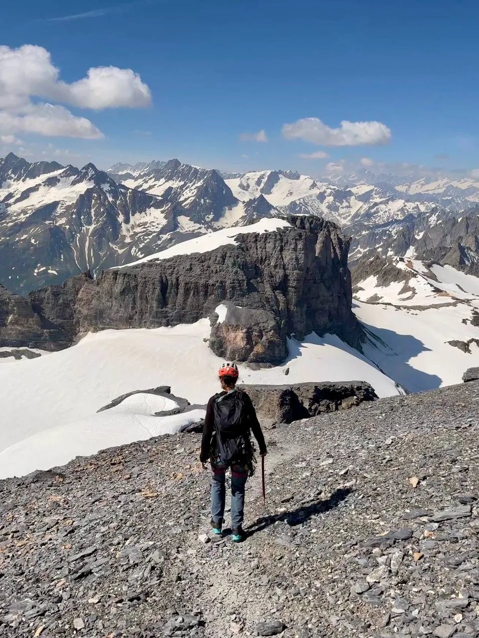 A hiker descending a scree path towards a vast panorama of snow-streaked Uri Alps peaks.