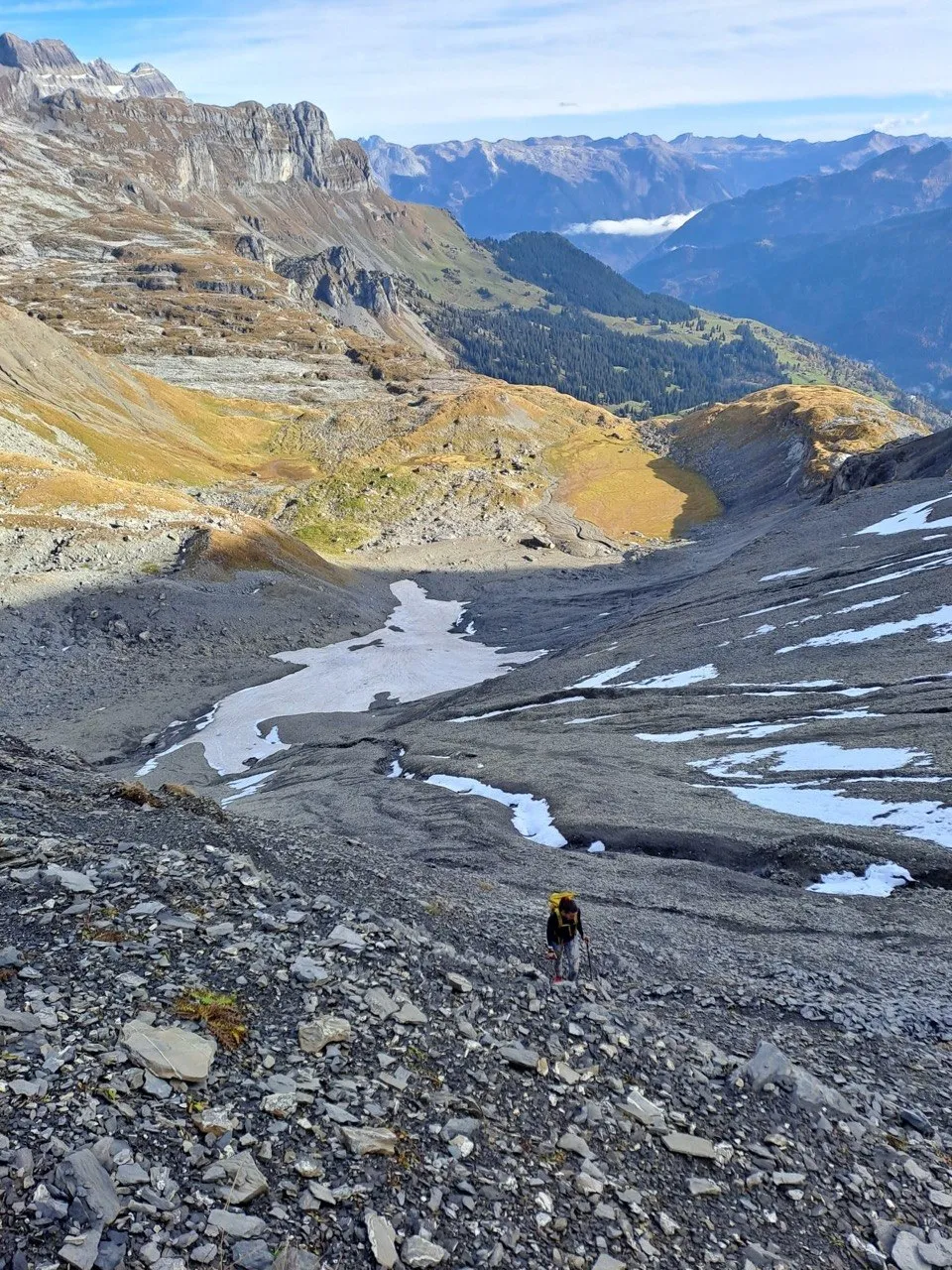 Hiker crossing scree and snow patches in a wide mountain basin.