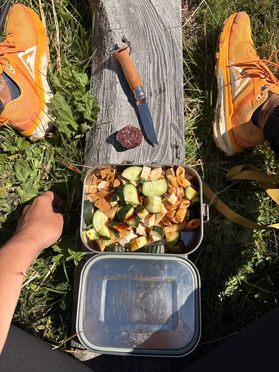 Pasta salad in a tin box on a weathered wooden bench, a pocket knife and a slice of salami beside it, orange trail shoes below.