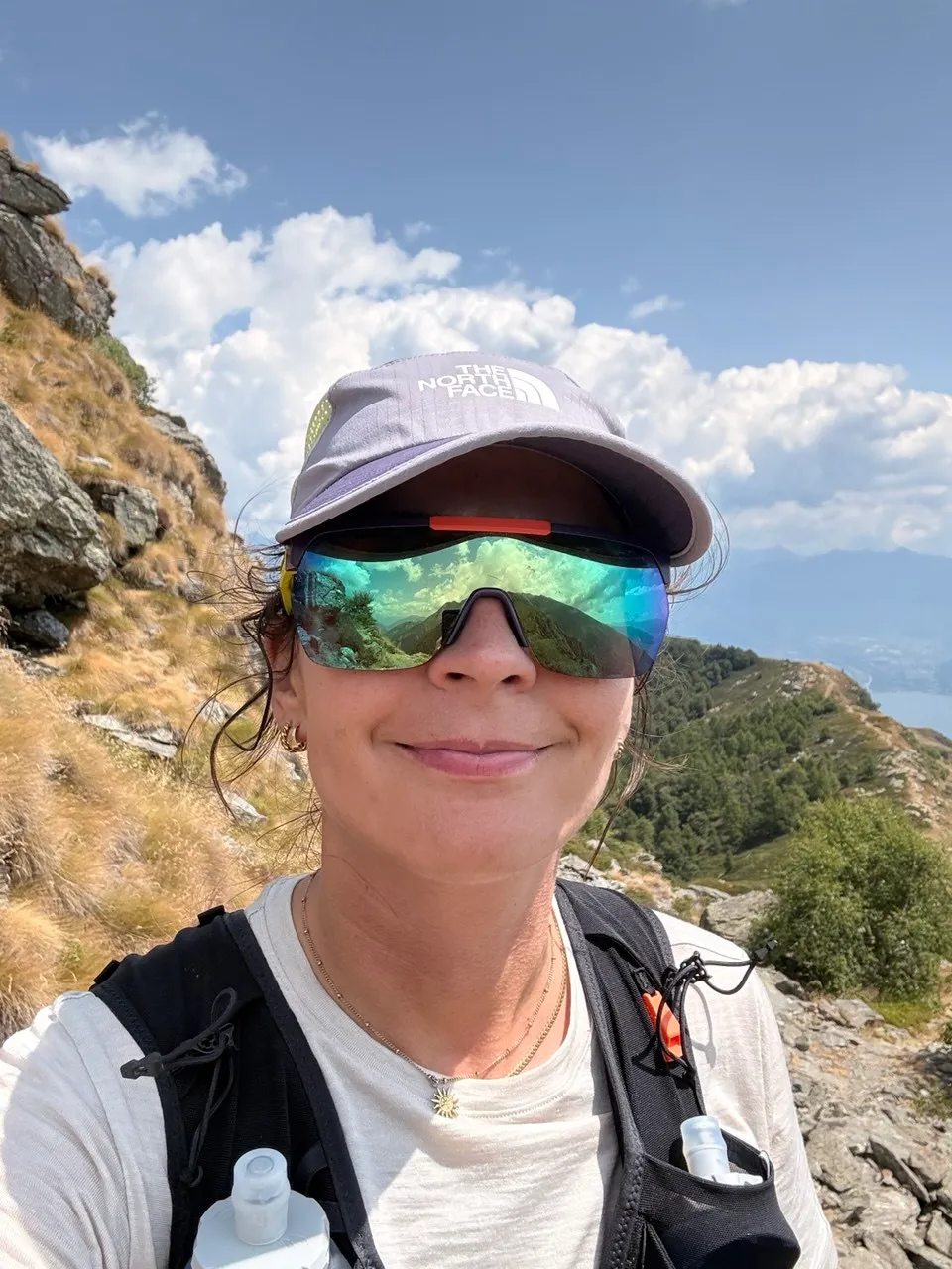 A smiling runner in a cap and mirrored sunglasses wearing a running vest with soft flasks, the ridge and the lake behind.