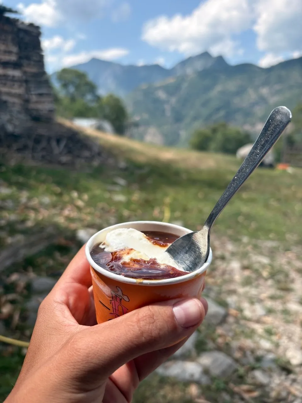 A hand holding a small orange paper cup of coffee topped with cream, an old stone ruin and mountains behind.