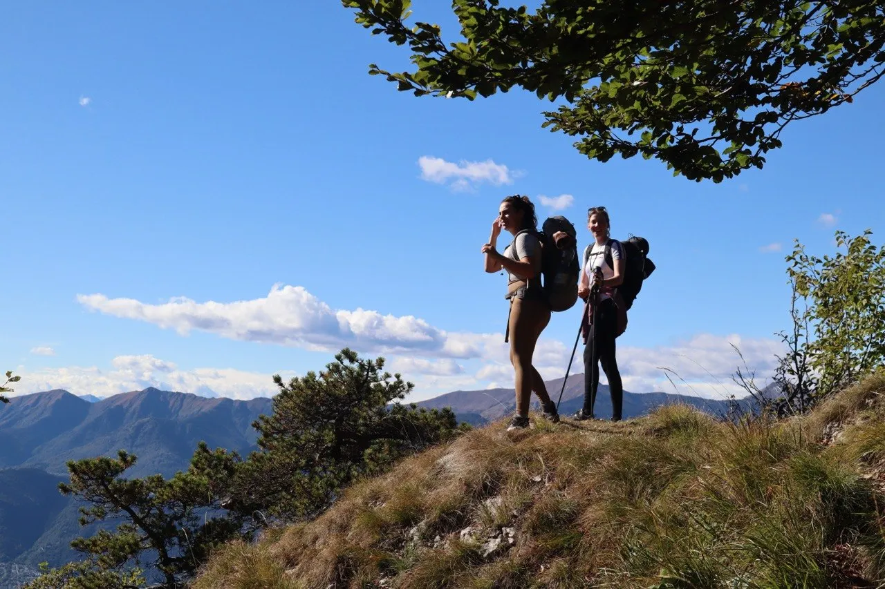 Two hikers with backpacks standing on a grassy ridge, mountains in the background.