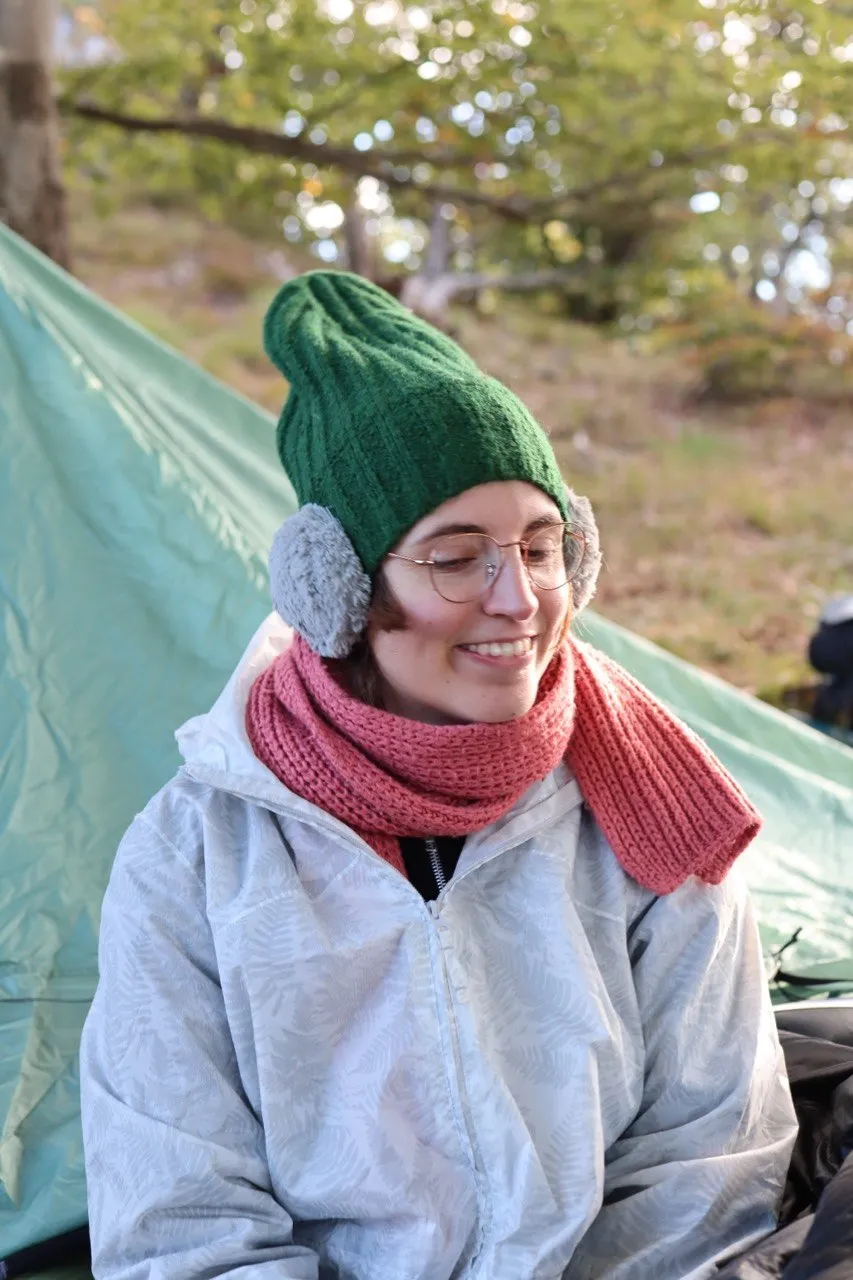 Portrait of a hiker bundled up in a green beanie, earmuffs, and pink scarf at camp.