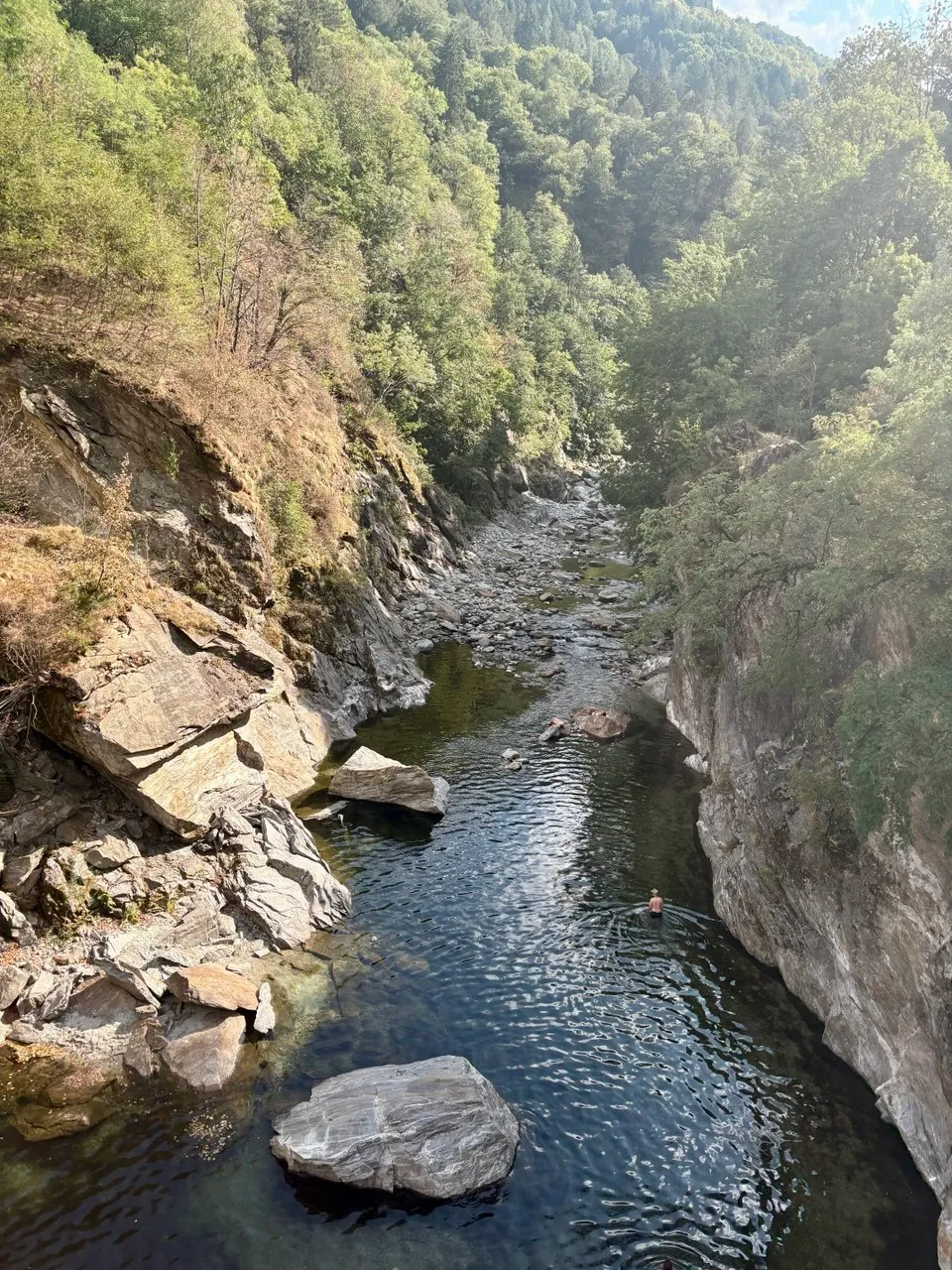 A narrow wooded river gorge from above, a swimmer in a deep clear pool among big smooth boulders.