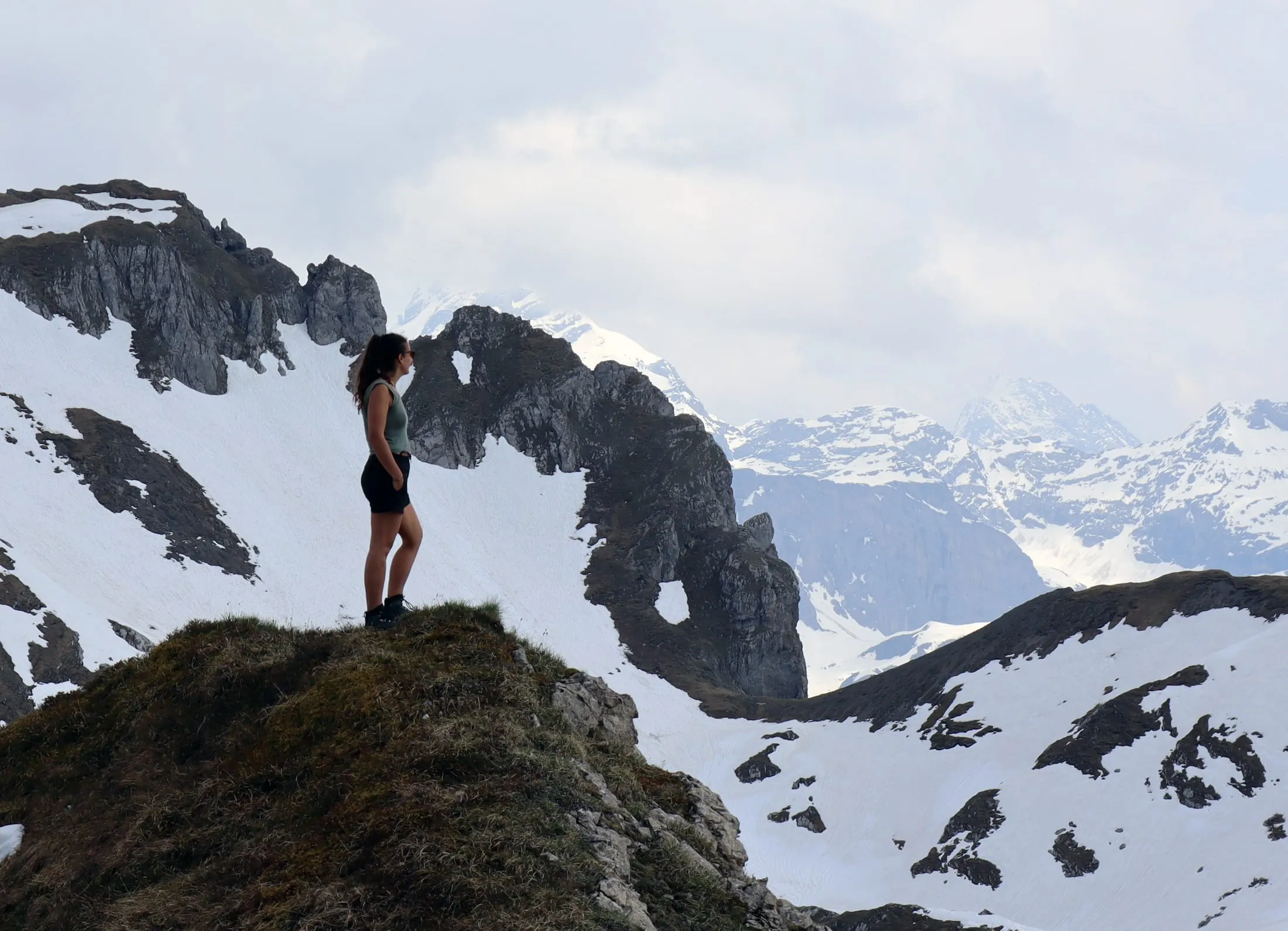Hiker in a green tank top standing on a grassy outcrop, snow-streaked peaks rising in the distance.