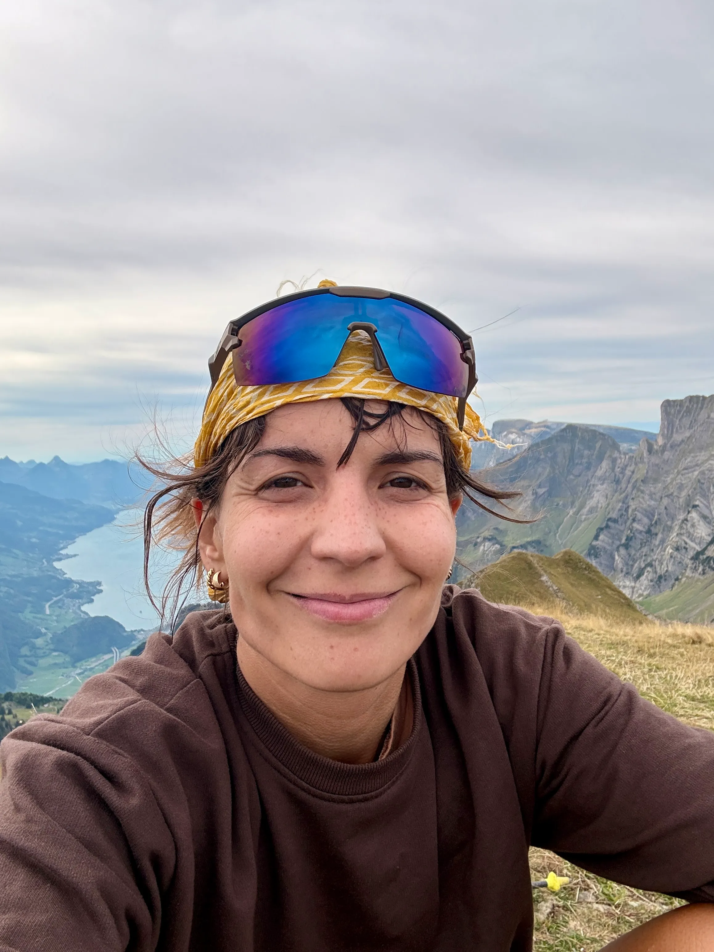 A summit selfie: a smiling hiker with a headscarf and sports sunglasses, the ridge and Lake Walen behind.