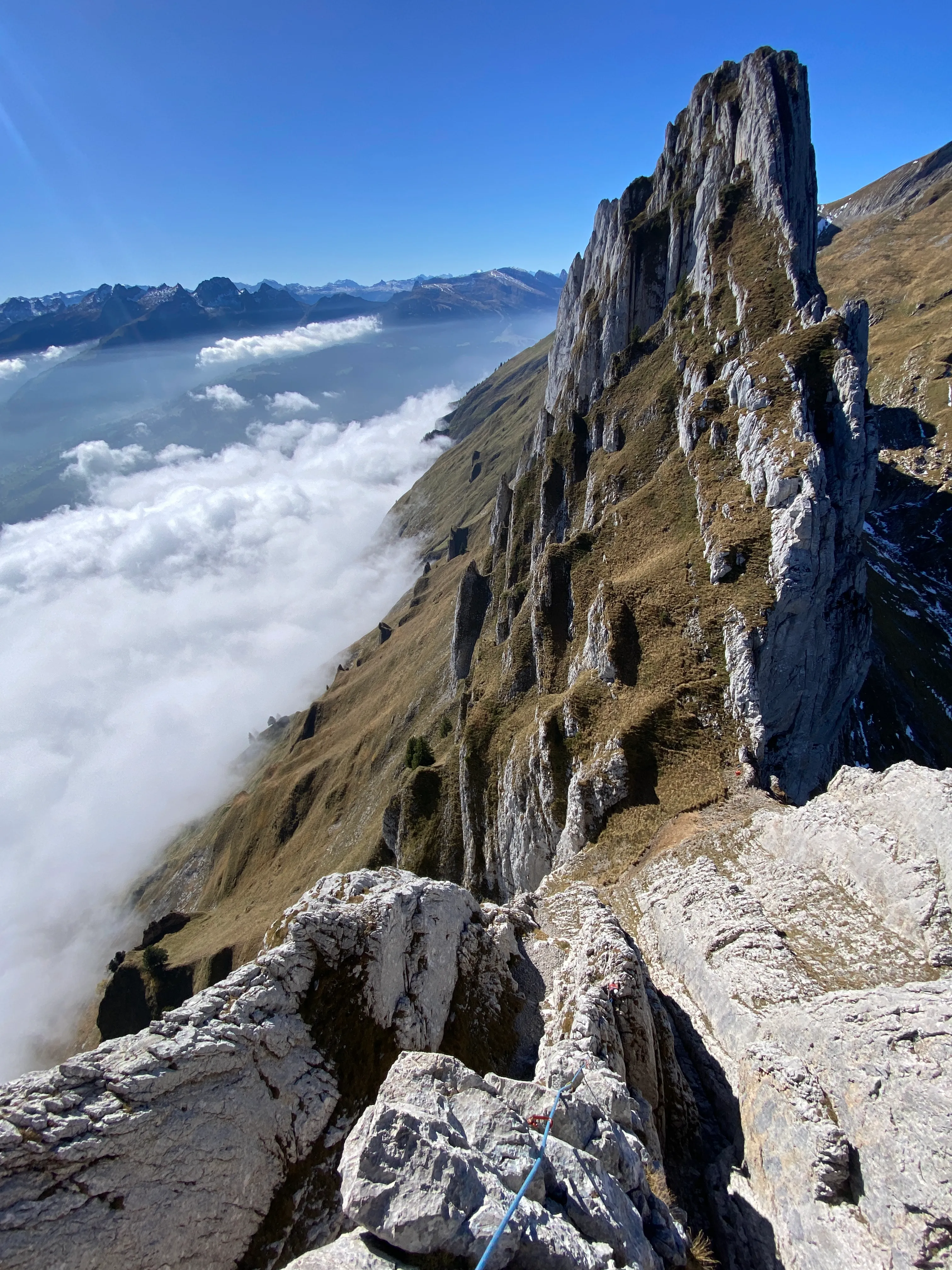 A serrated limestone wall dropping into a sea of fog, distant alps beyond under a blue sky, a climbing rope in the foreground.