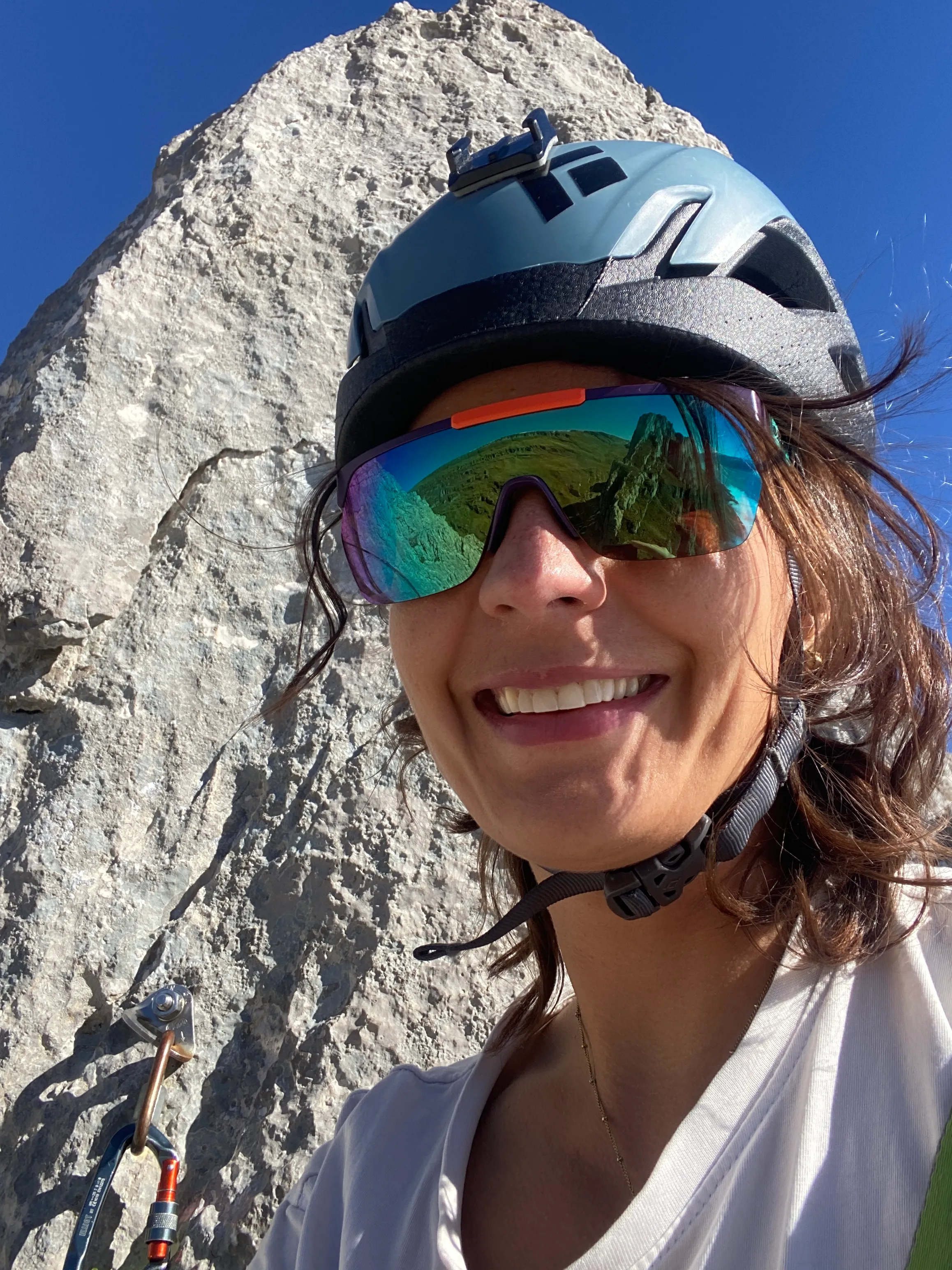 Selfie of a laughing climber in helmet and sunglasses below a pale rock tower under a blue sky.