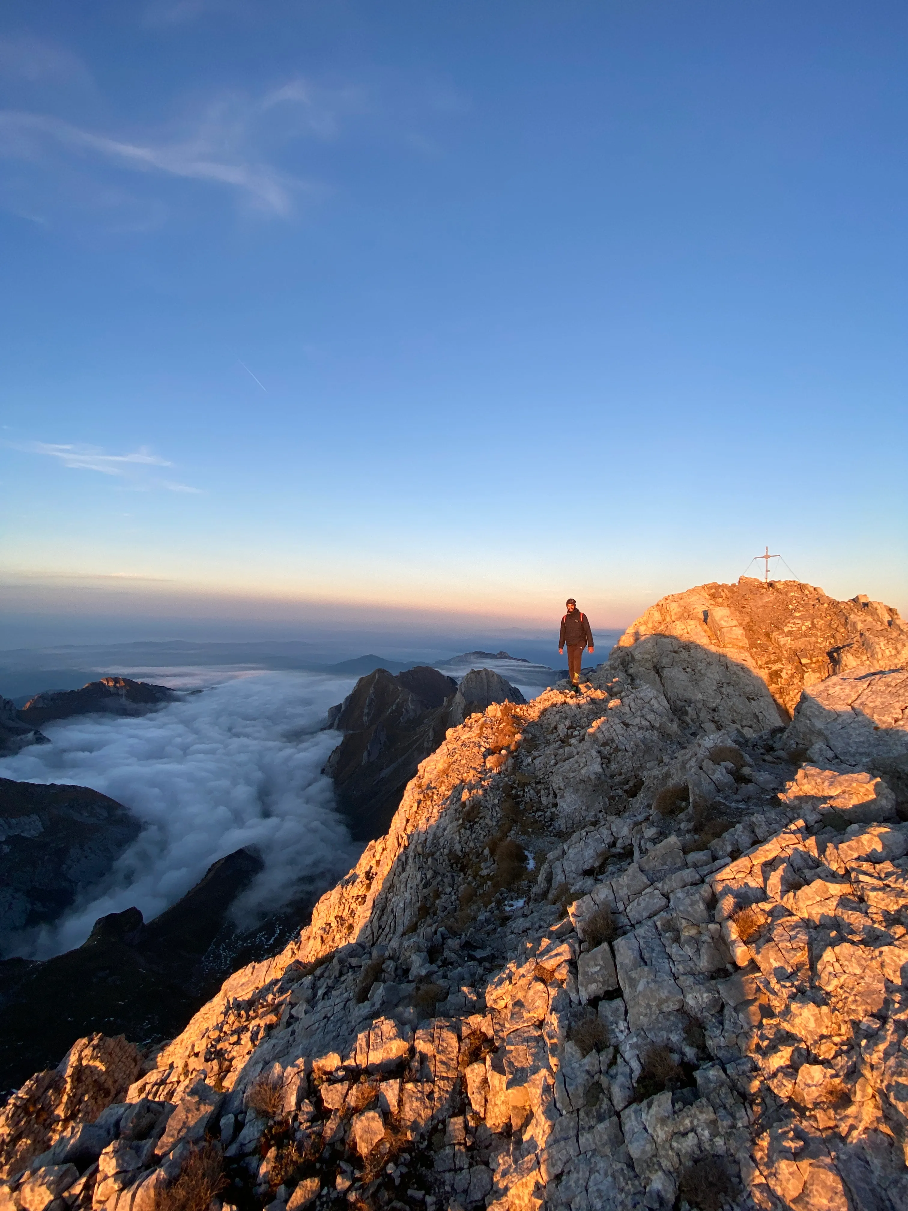 A hiker on the summit ridge by the Altmann summit cross in the last evening light, a sea of fog below.