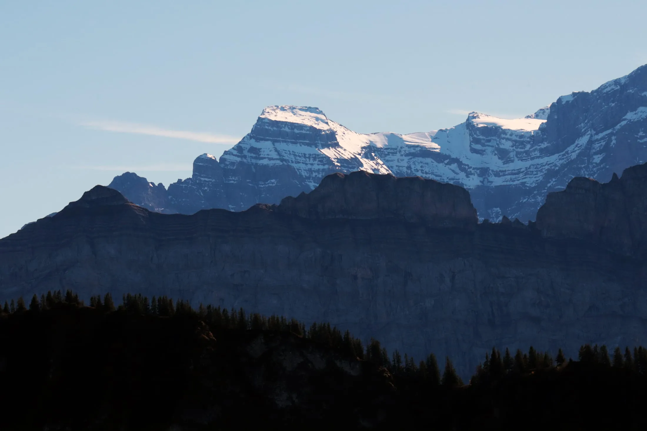 Layered Glarner limestone peak topped with snow above a dark rock wall in backlight.