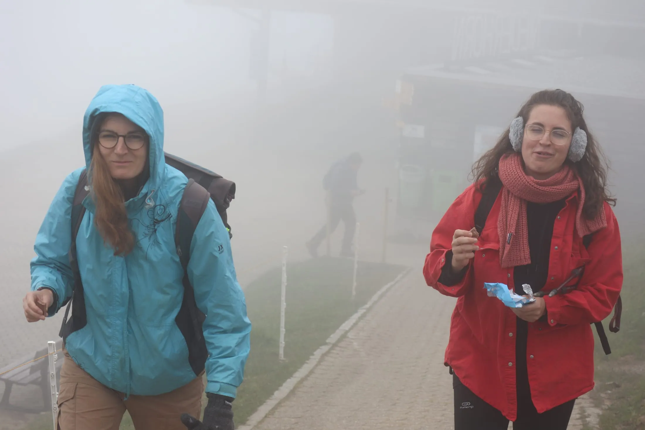 Two hikers bundled up in thick fog at the Niederhorn station eating chocolate.