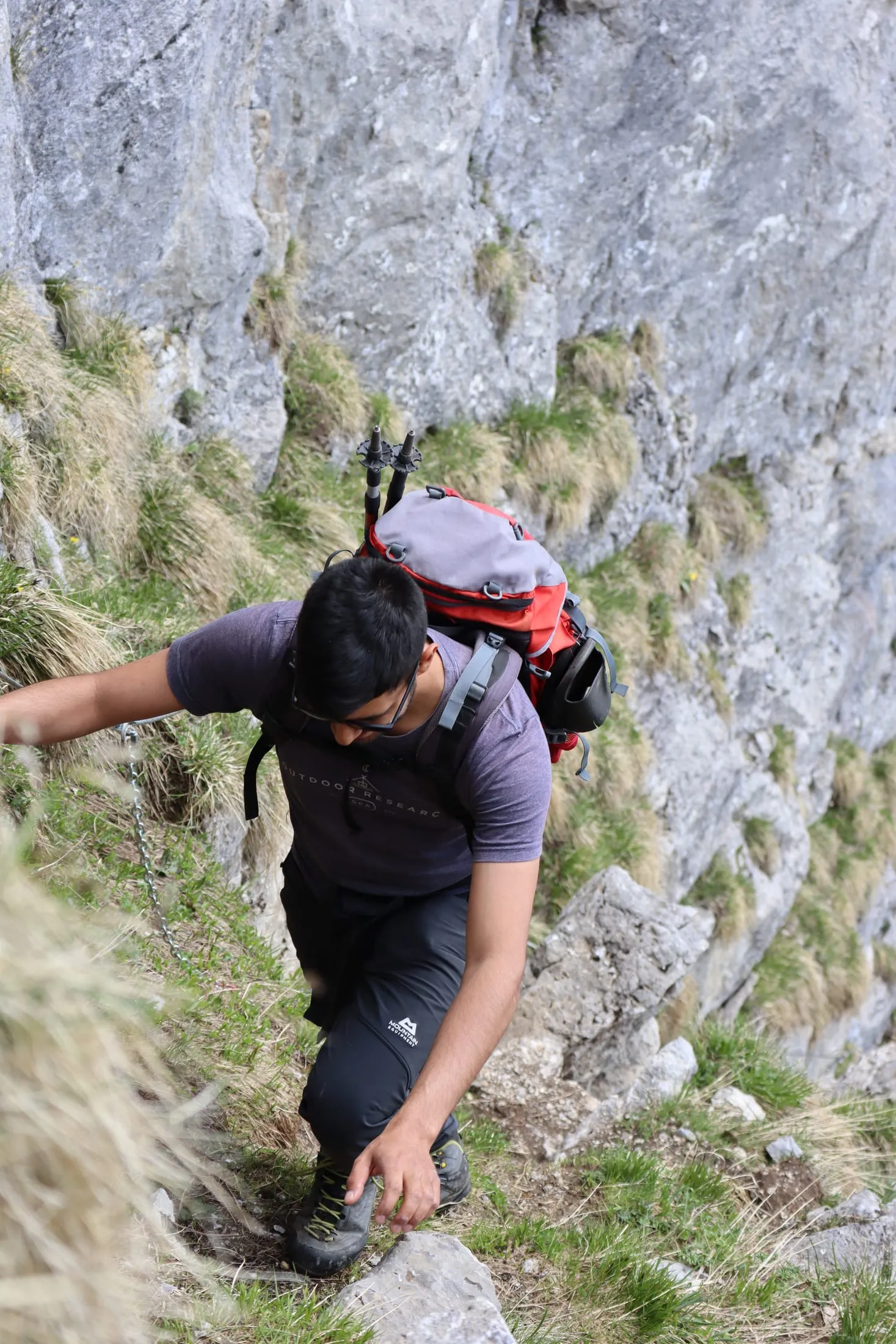 Looking down on a hiker clinging to grassy ledges between slabs of grey rock.