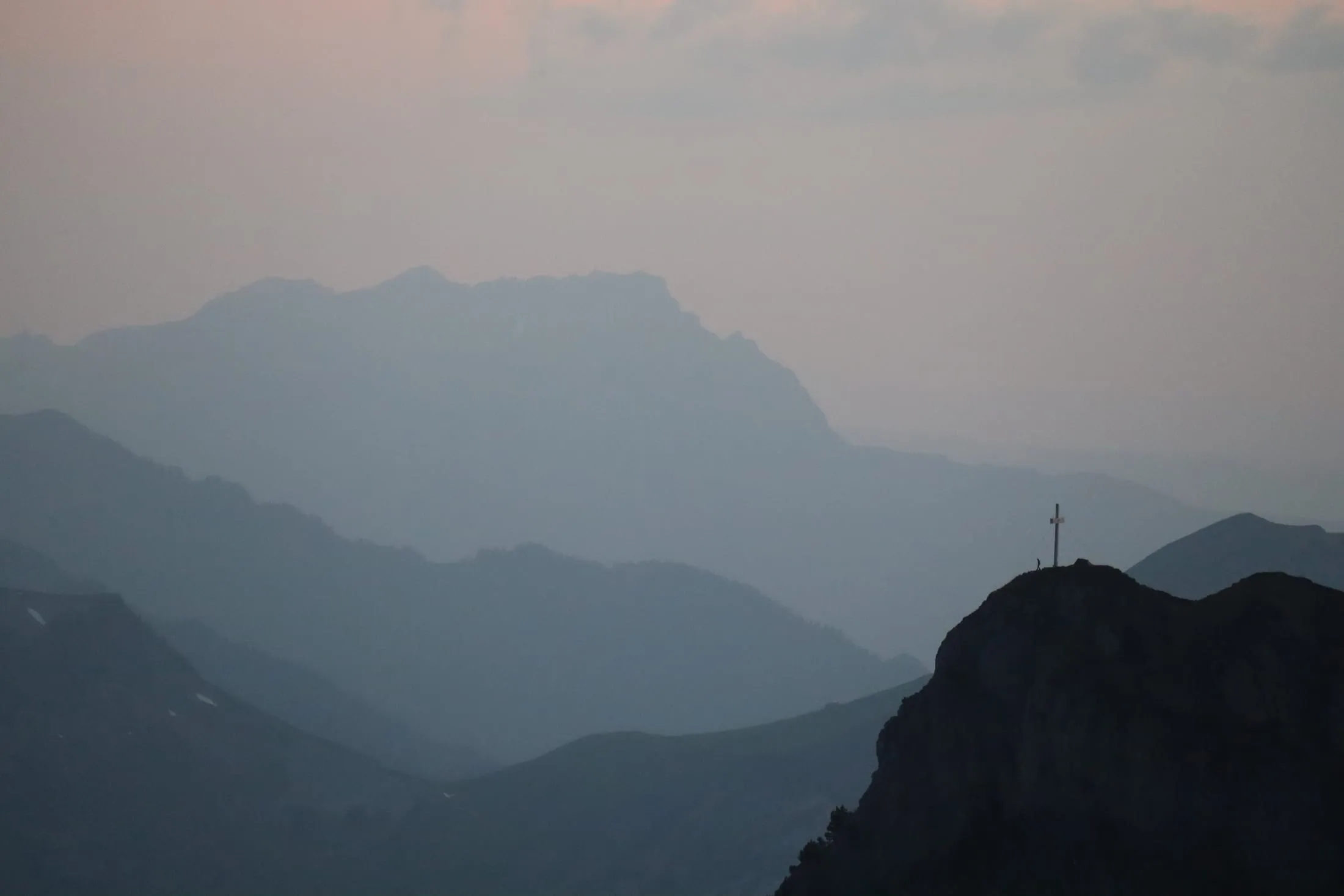 Tiny figure beside a summit cross with layered hazy ridges receding into dusk.