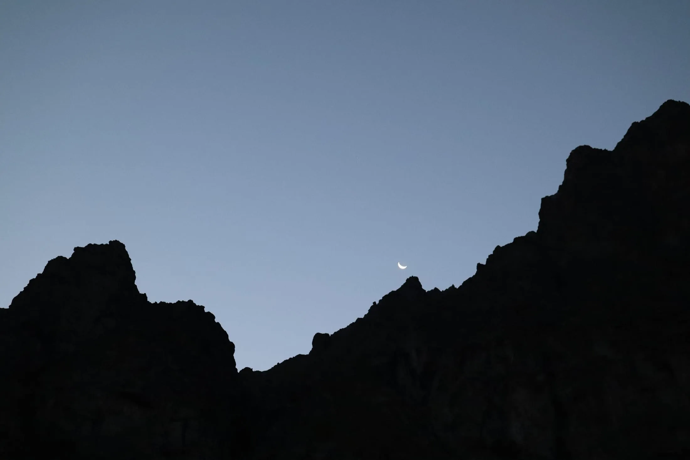 Thin crescent moon between two silhouetted rocky summits at dusk.