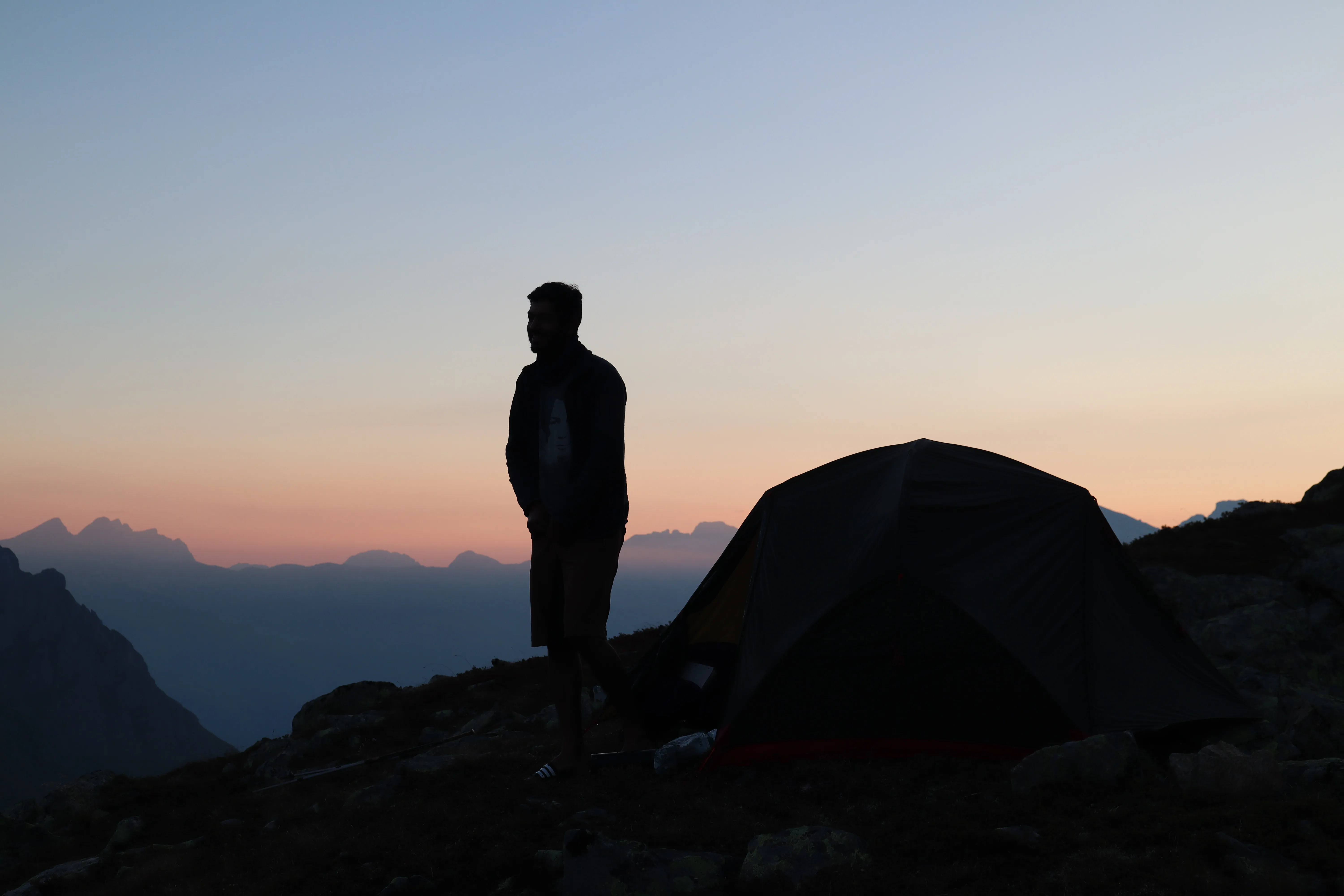Silhouette of a person next to a tent against a pink-orange evening sky over the valley.