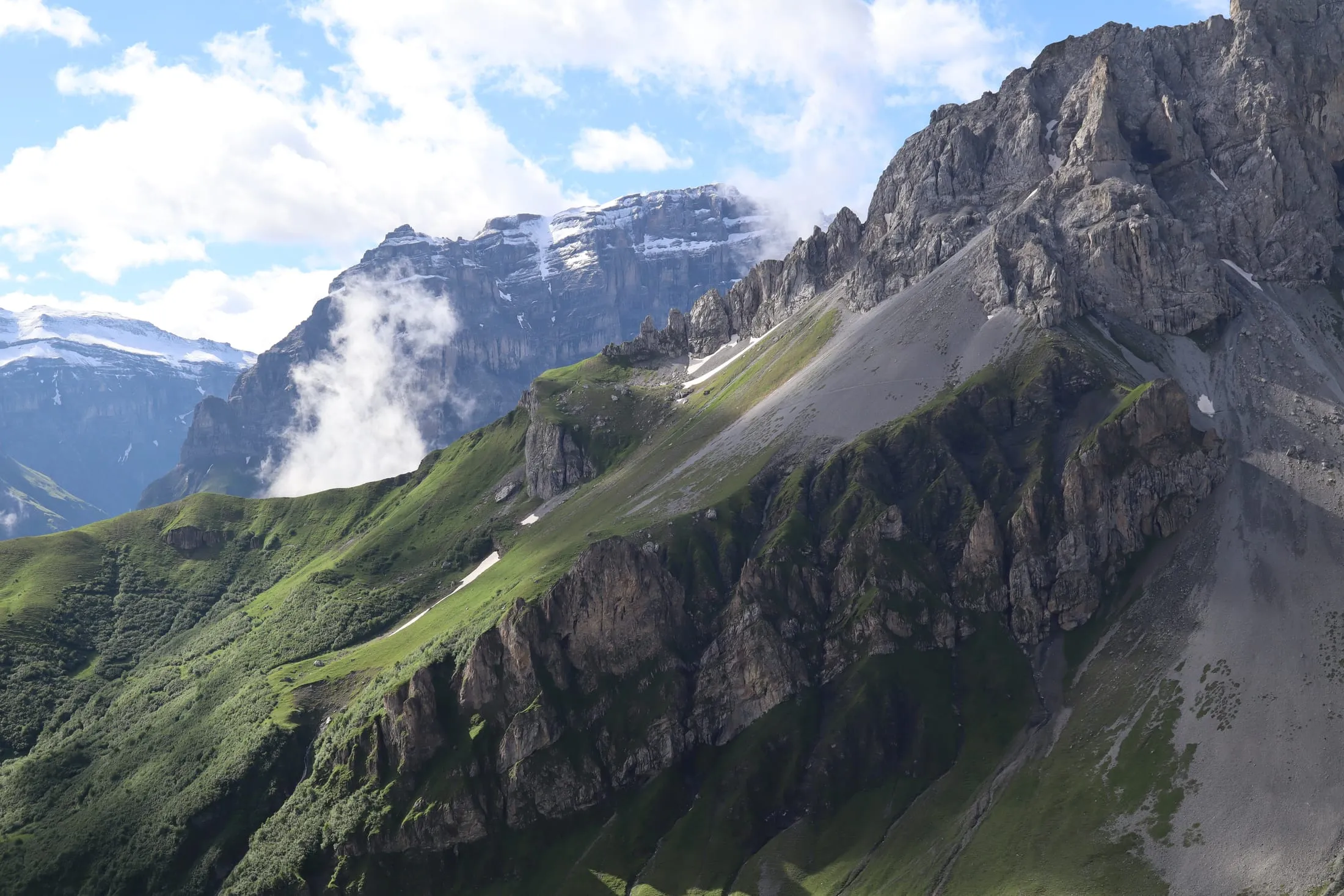 Rugged rock flanks with green slopes and wisps of cloud, snow-capped peaks behind.