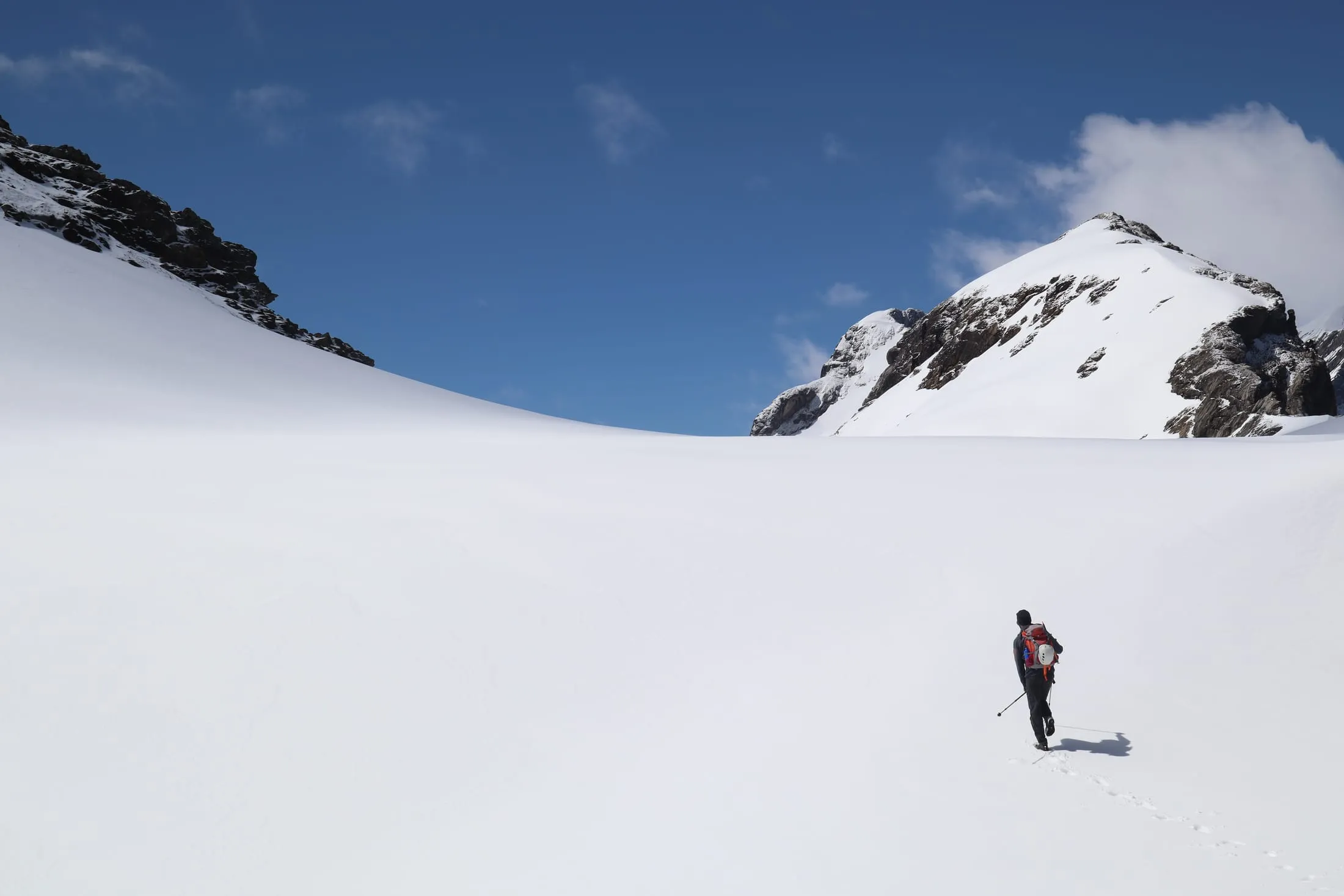 A lone mountaineer crosses a vast snowfield toward a snowy peak under a deep blue sky.