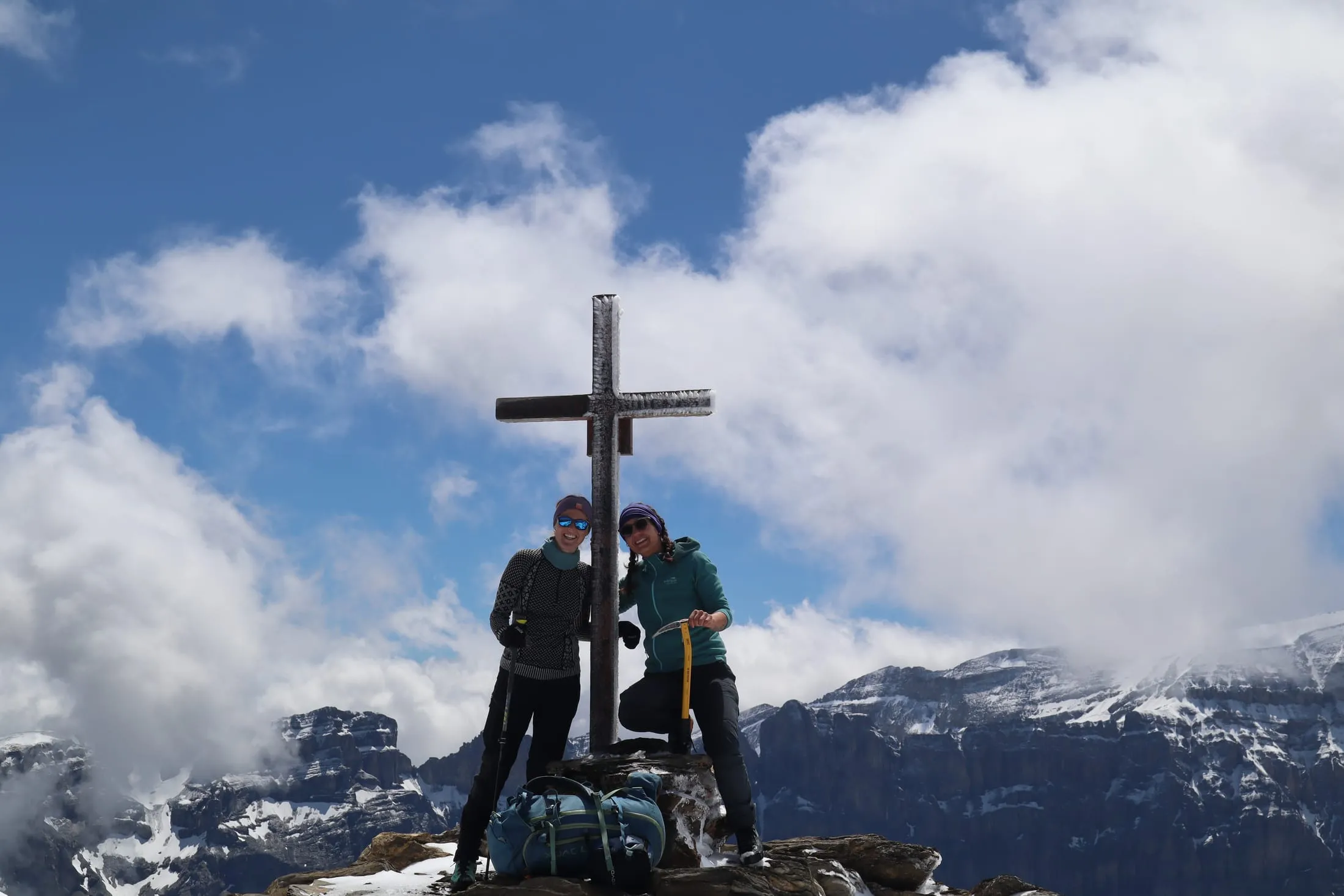 Two mountaineers stand with a rope at the snowy summit cross before clouds and distant peaks.