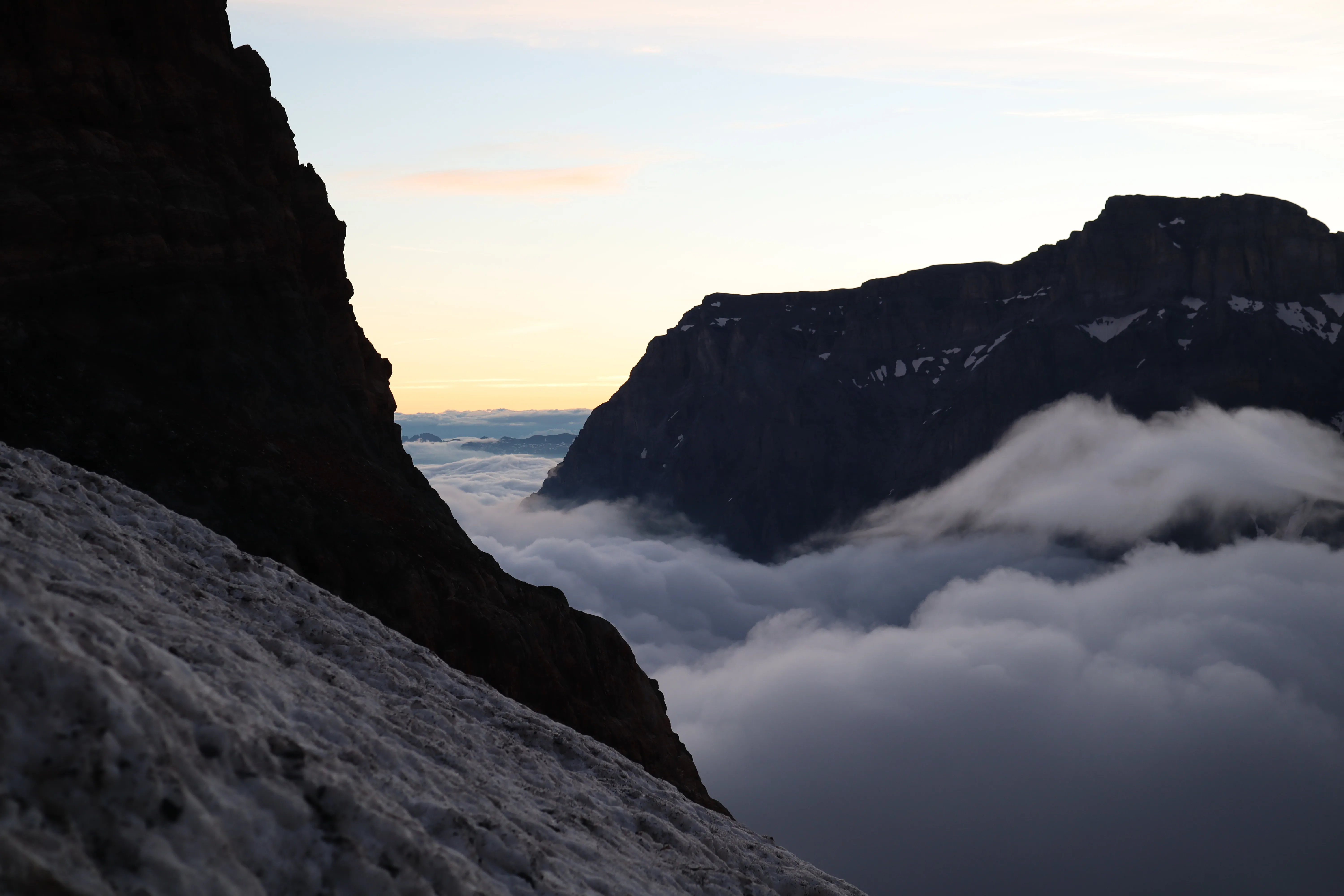 Dark peak silhouettes above a sea of clouds at dawn, an orange band along the horizon.