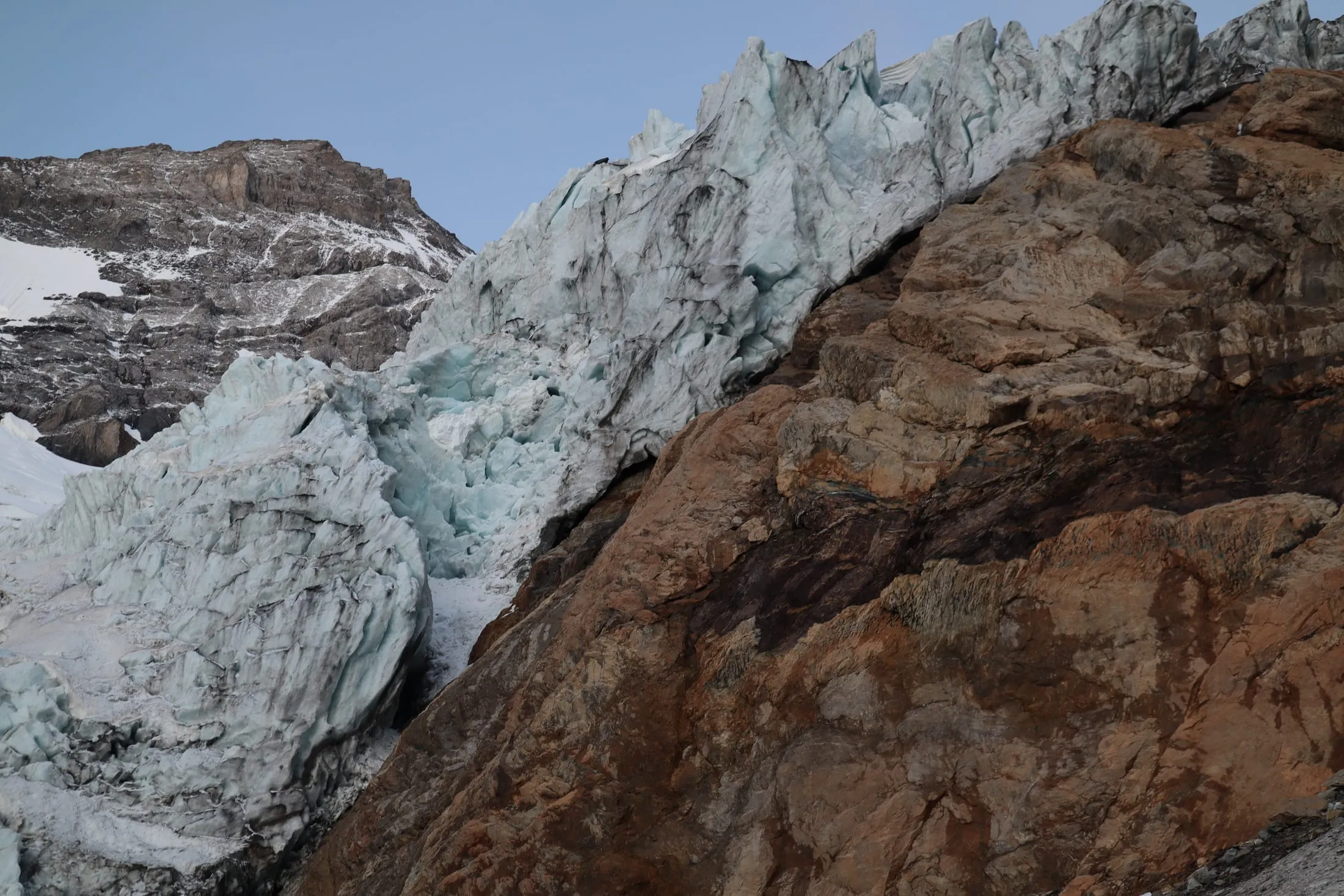Blue glacier tongue with crevasses beside a brown rock wall in the early morning light.