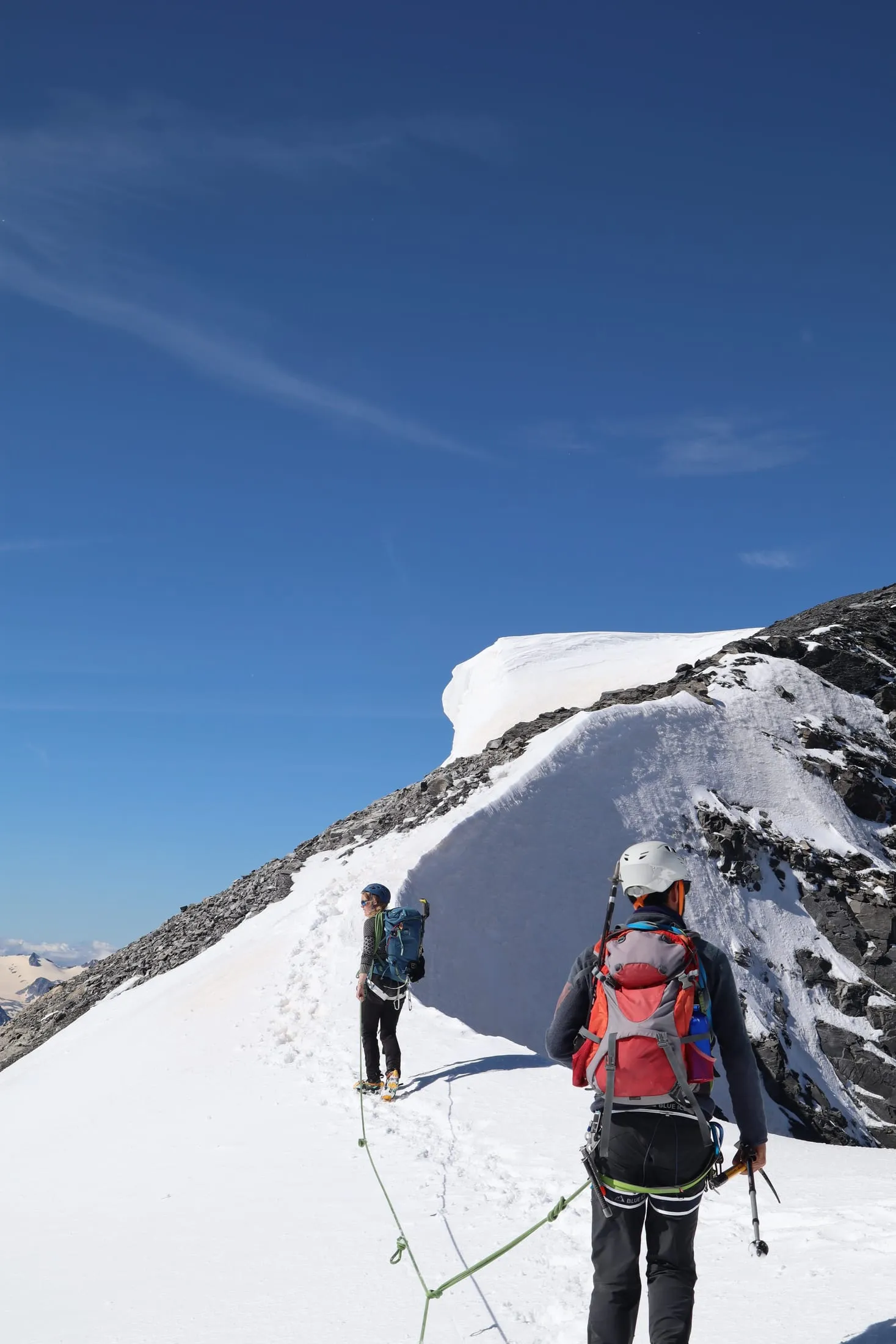 Two roped mountaineers climb a snowy ridge under a deep blue sky.