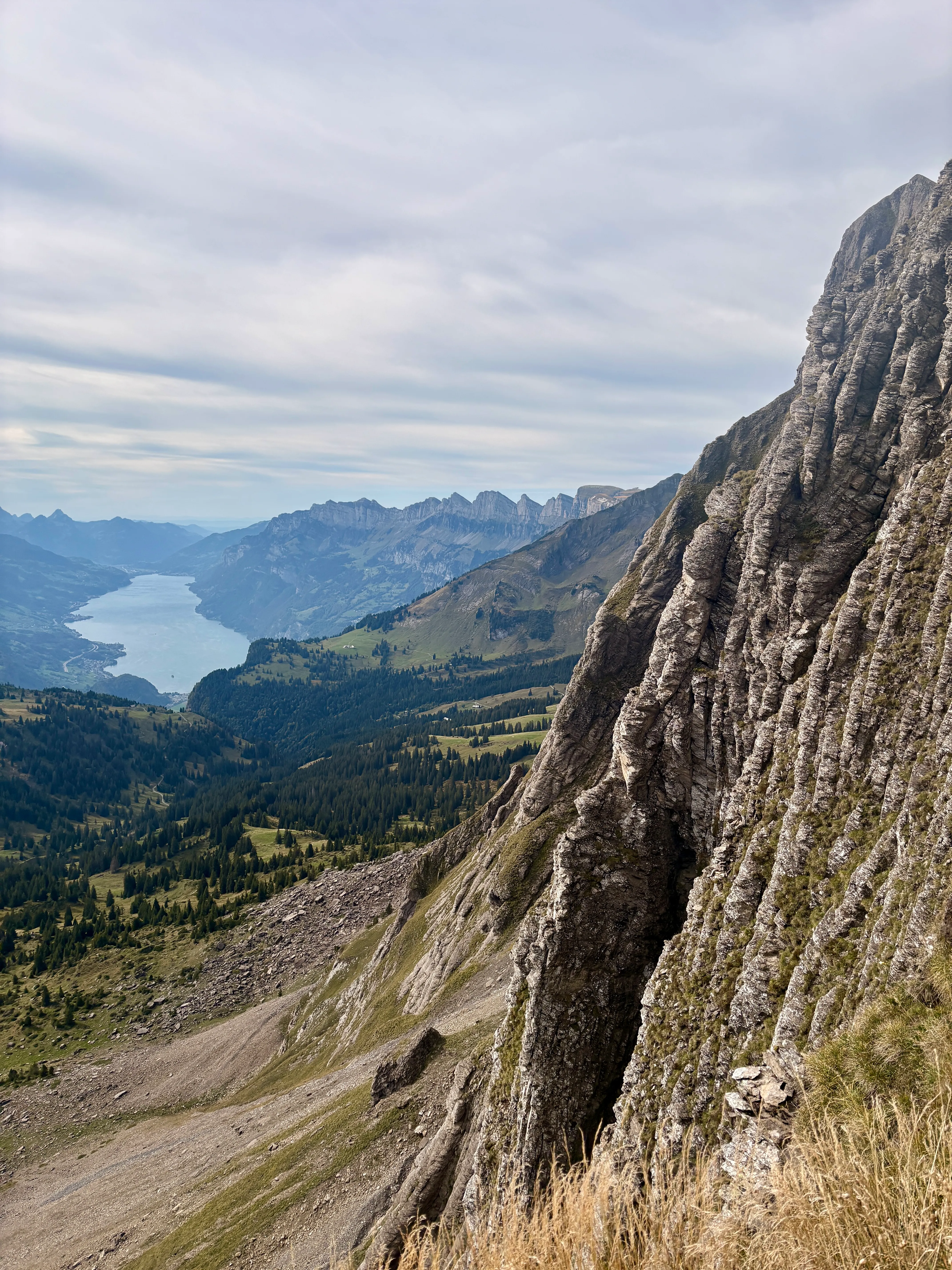 A steep layered limestone wall with a view down to Lake Walen and the Churfirsten.