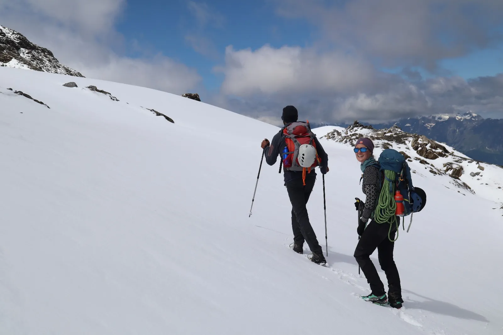 Two mountaineers cross a broad snowfield, the woman turning back with a smile, rope coiled on her pack.