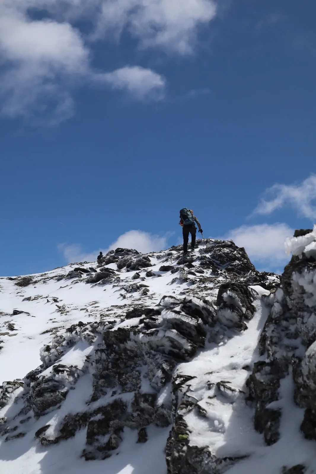 A mountaineer climbs a snow-dusted rocky ridge toward the summit under a blue sky.
