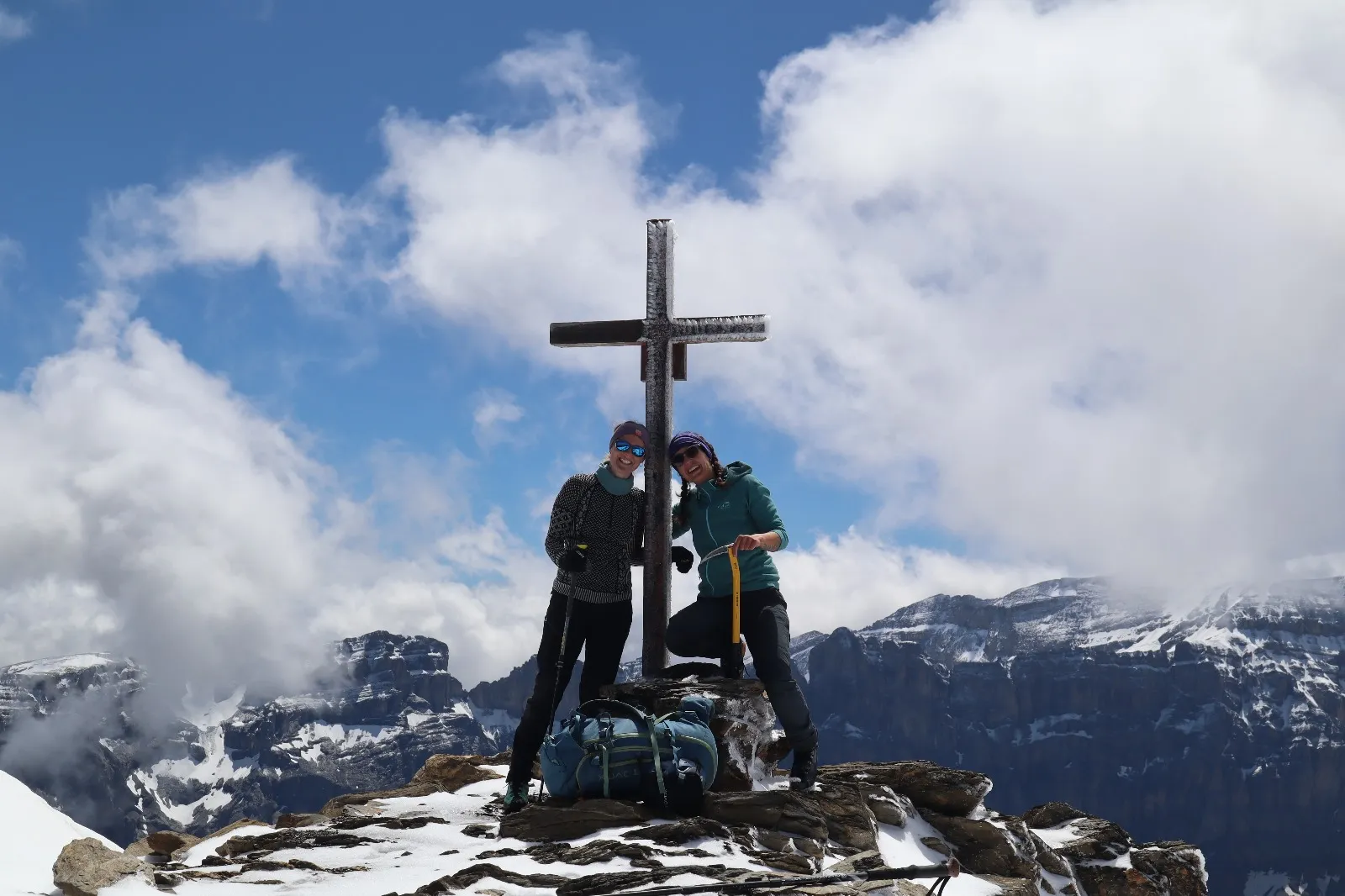 Two hikers at the summit cross with cloud-wrapped mountain ranges behind.