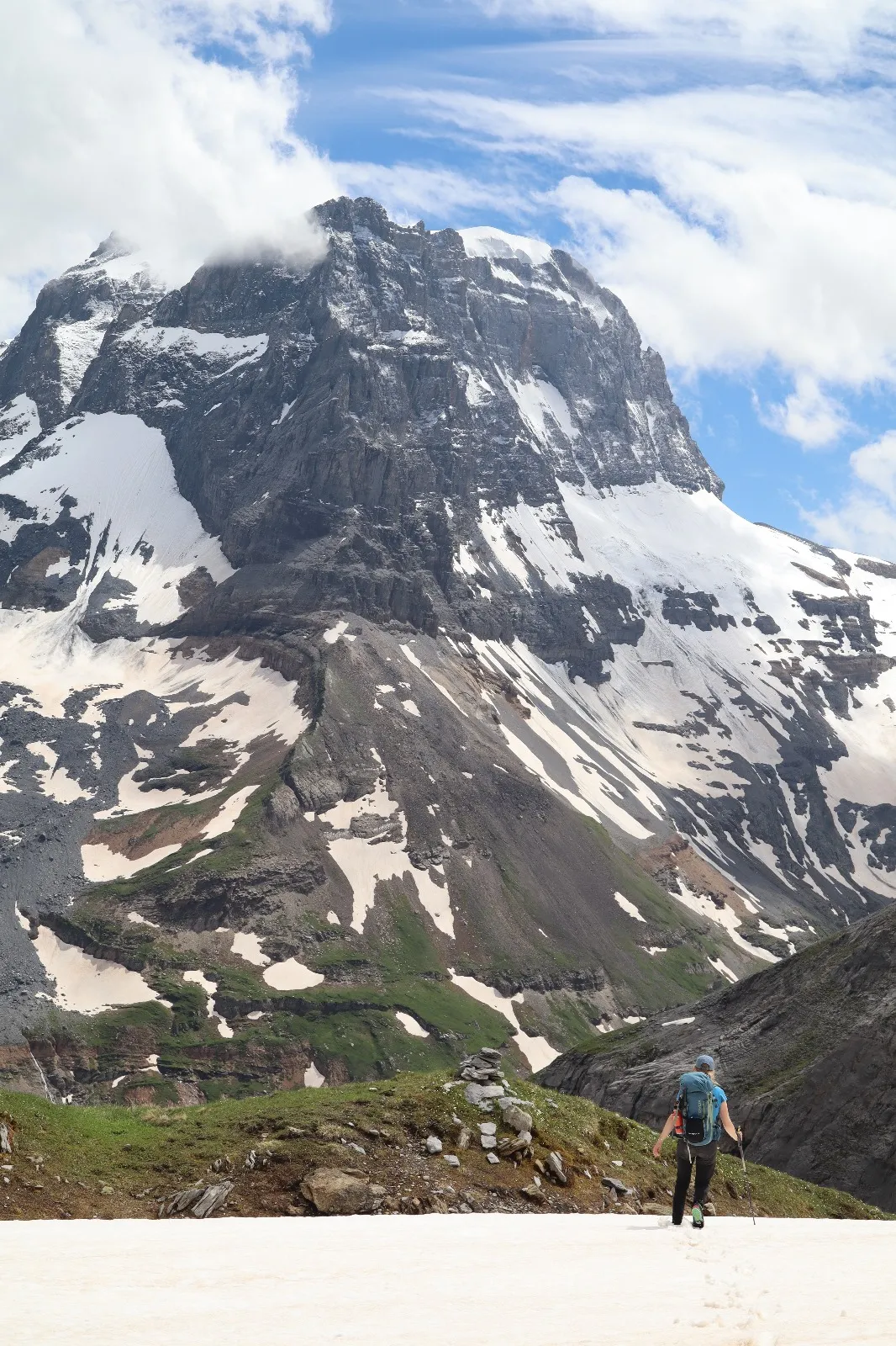 A hiker crosses a snowfield toward the dark, snow-streaked rock pyramid of the Gemsfairenstock.