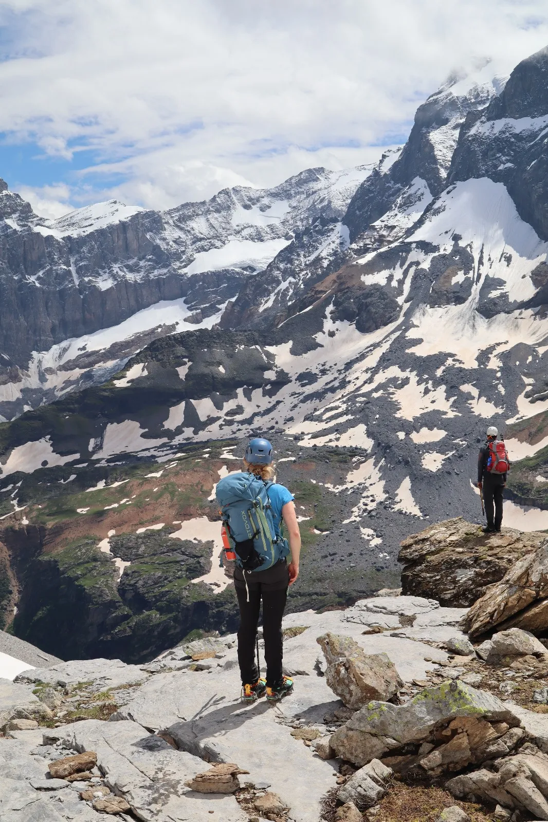A helmeted hiker stands on a rock slab looking out over a snow-filled cirque.
