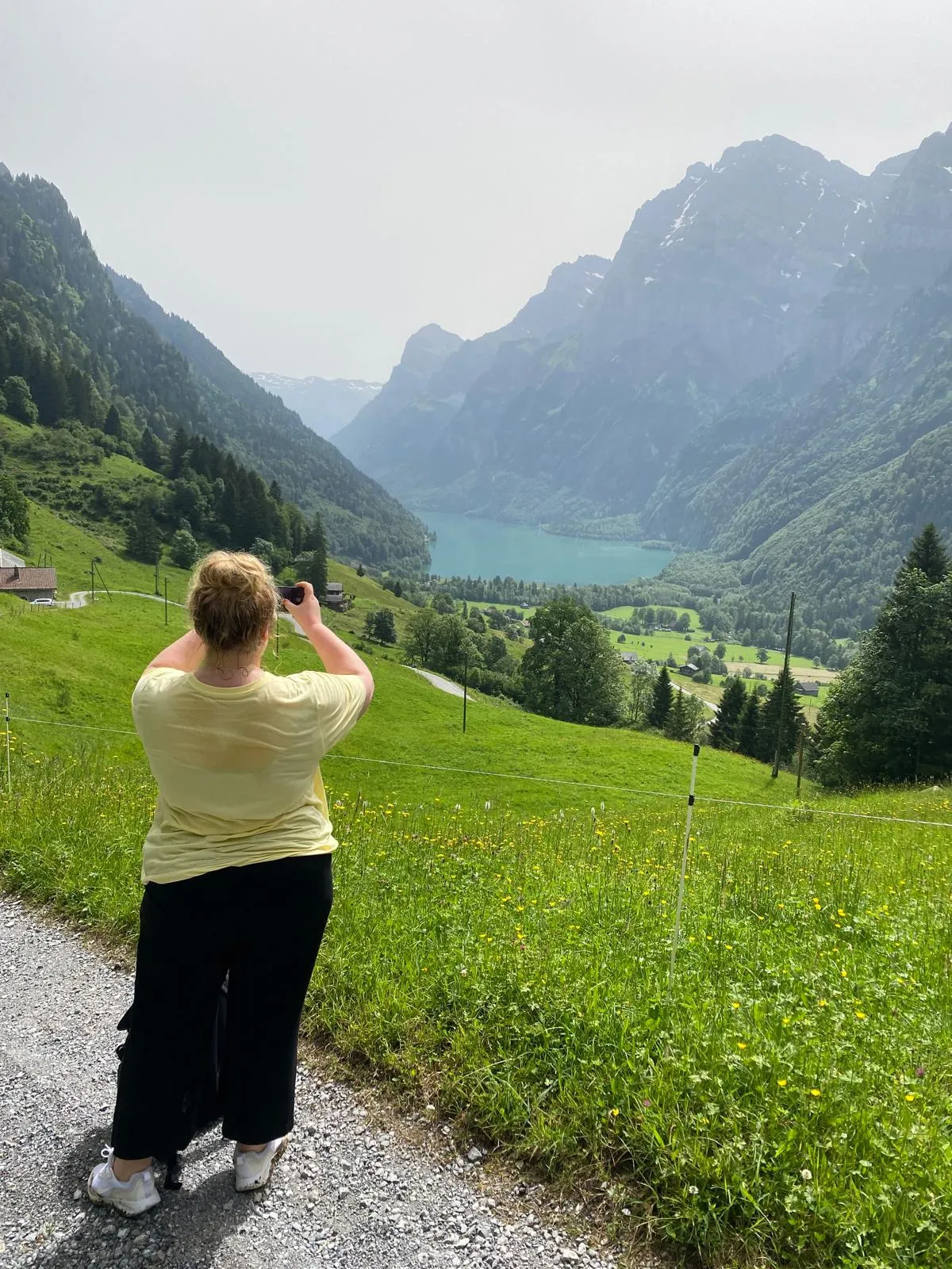 Hiker in a yellow shirt photographing the turquoise lake from a wildflower meadow.
