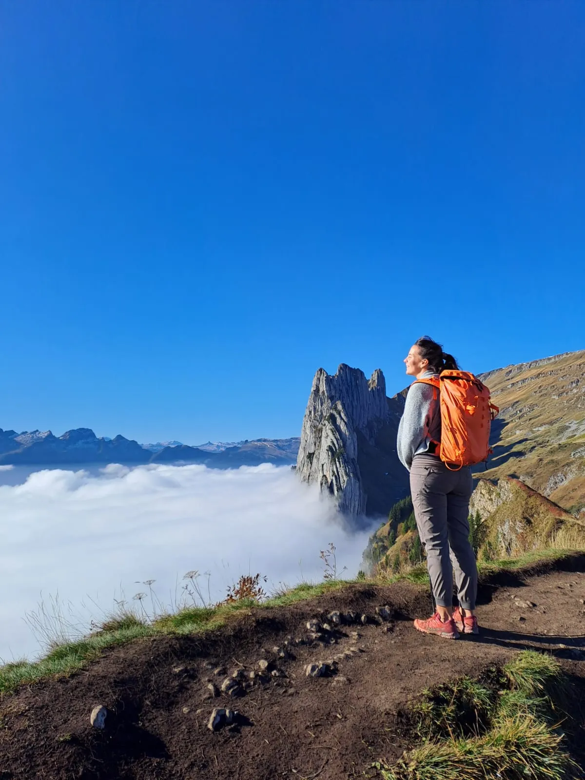 A hiker with an orange backpack looks out over a sea of fog toward the rock spire of the Saxerlücke.