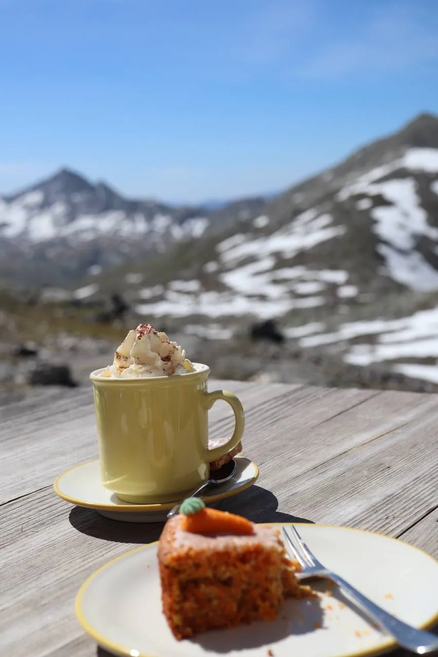 A cappuccino and a slice of carrot cake on a wooden hut terrace with snowy peaks behind.
