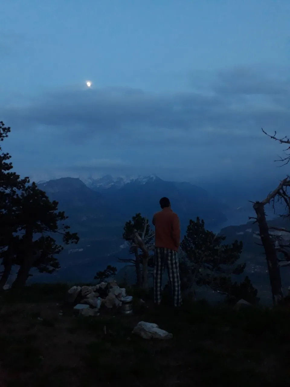 A figure in a fleece stands at blue hour looking out to shadowed peaks, the moon rising through cloud above the valley.