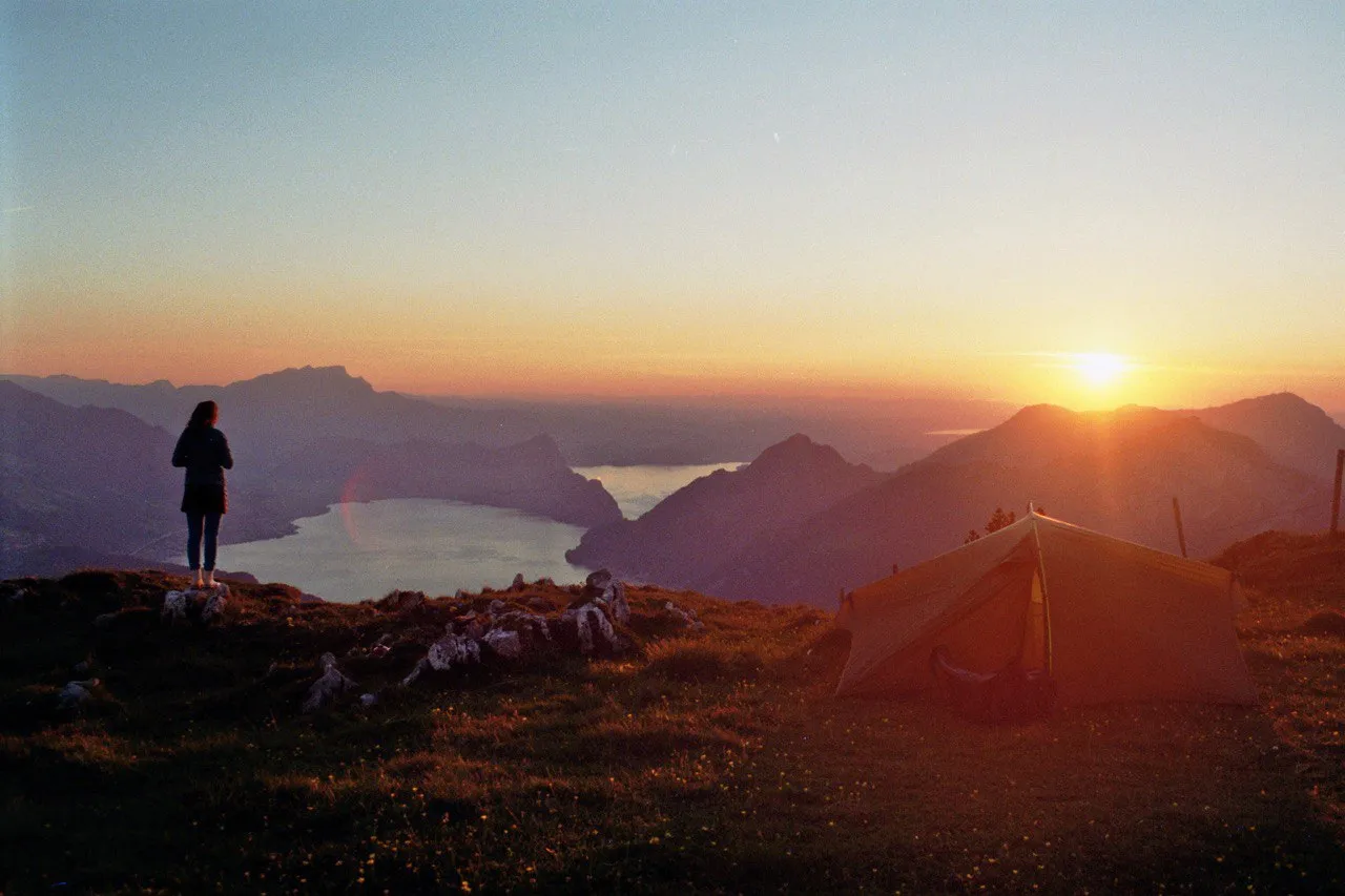 Tarp tent pitched on a grassy ridge at sunset, a hiker gazing over Lake Lucerne far below with the sun setting behind distant peaks.